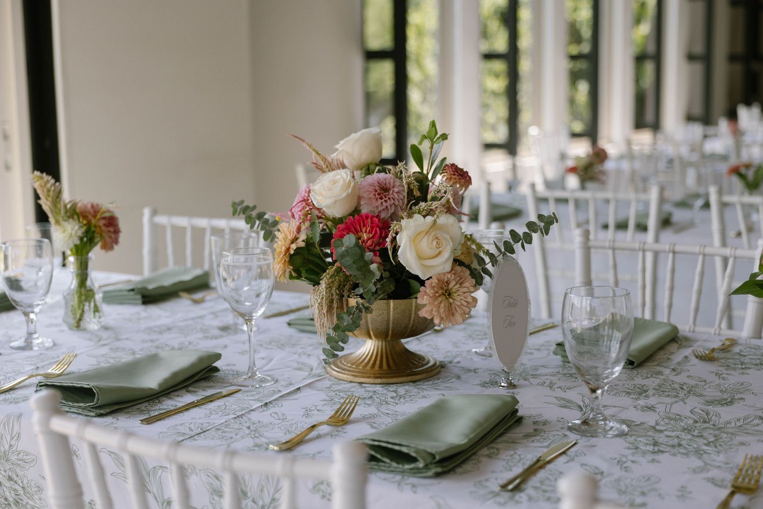 Elegant wedding reception table with gold floral centerpiece, green napkins, gold cutlery, and white chiavari chairs.