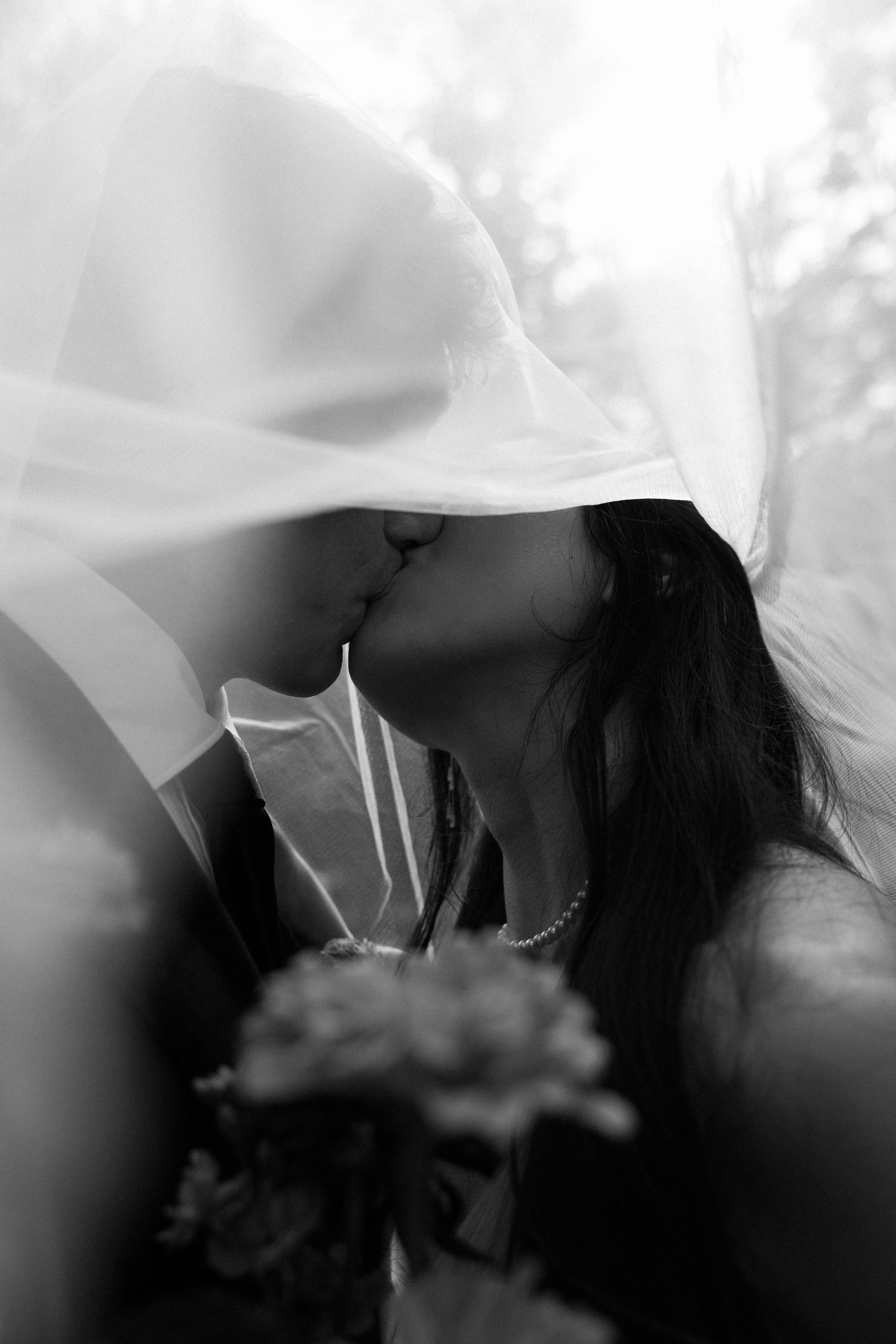 Black and white photo of a bride and groom sharing a kiss under a wedding veil, with a bouquet of flowers.