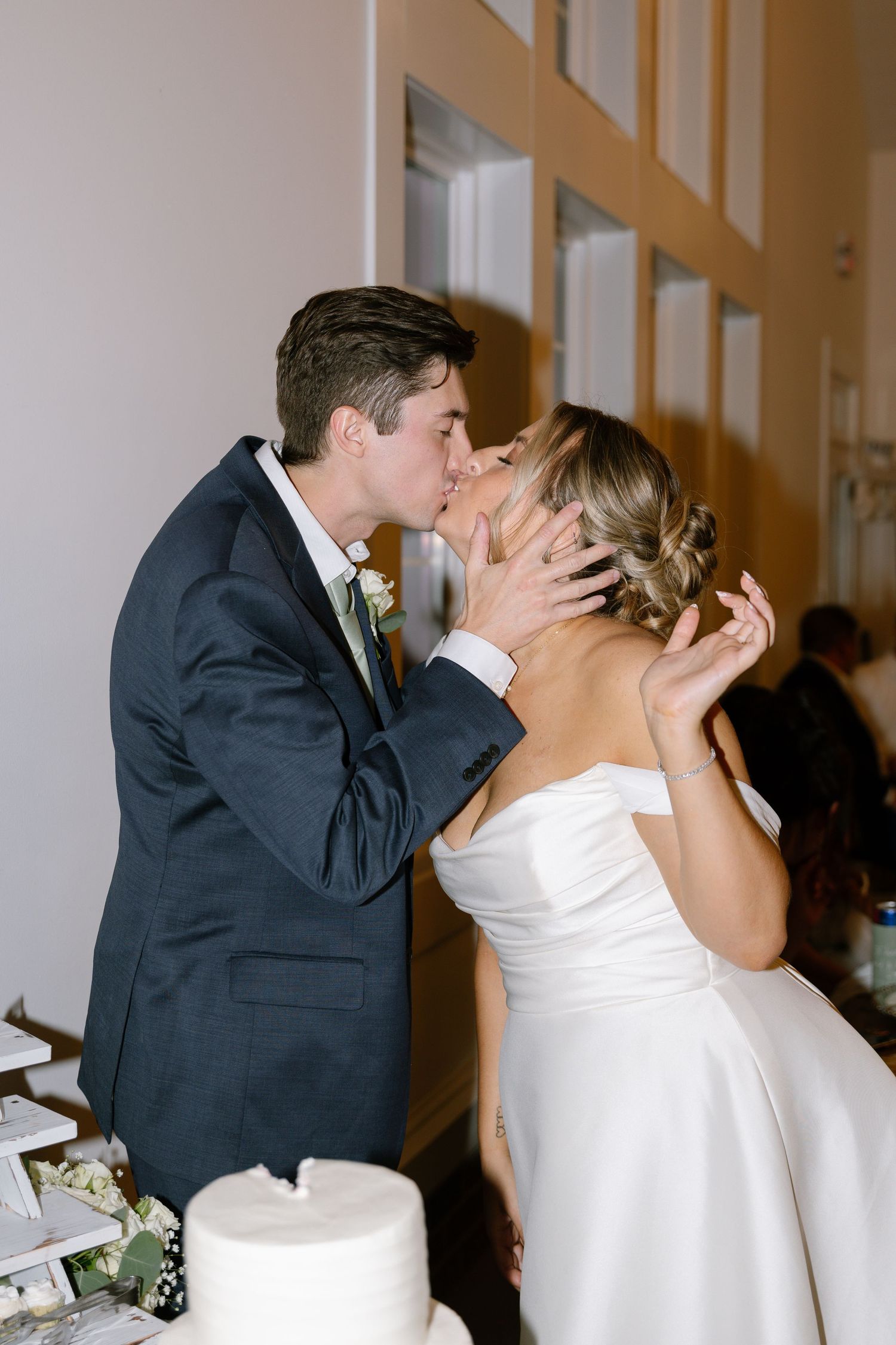 Groom tenderly cups bride's face for a romantic kiss beside their white wedding cake at the reception.