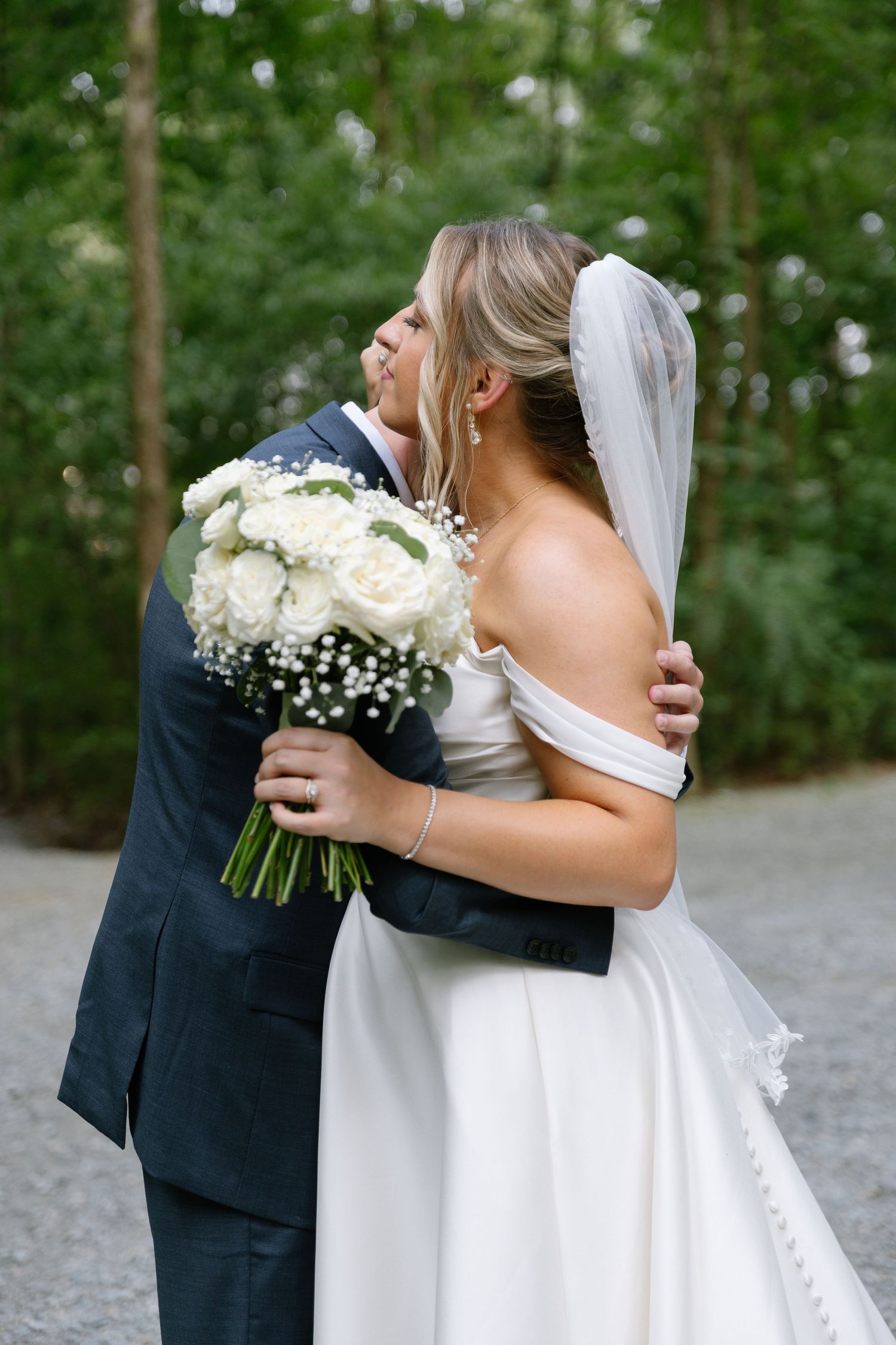 Bride and groom embracing outdoors, bride holding white rose bouquet with baby's breath, wearing veil and off-shoulder gown.