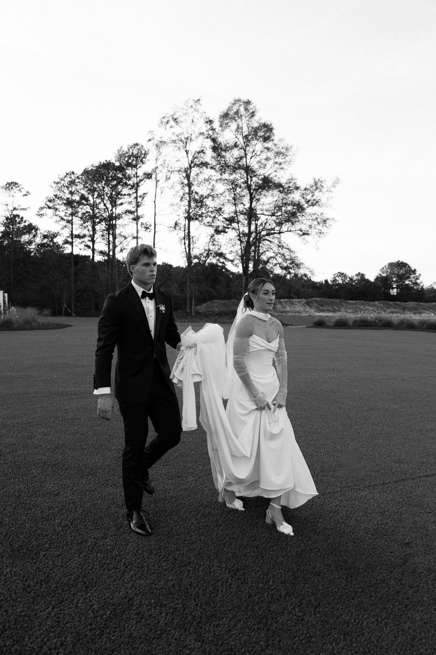 Bride and groom walking across an open field at dusk in a classic black and white wedding portrait.
