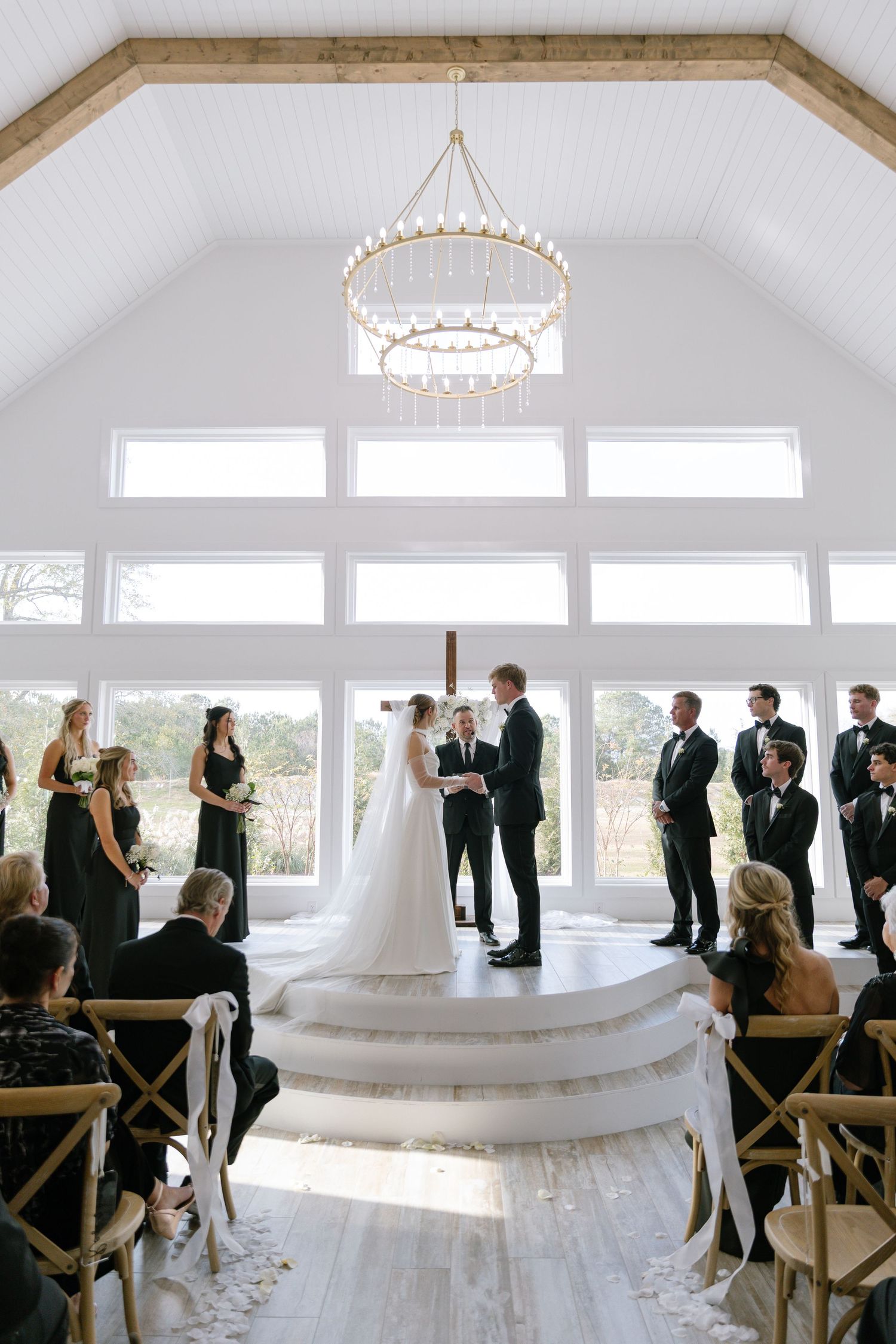 Elegant indoor wedding ceremony with bride and groom at altar beneath gold chandelier with black-clad wedding party.