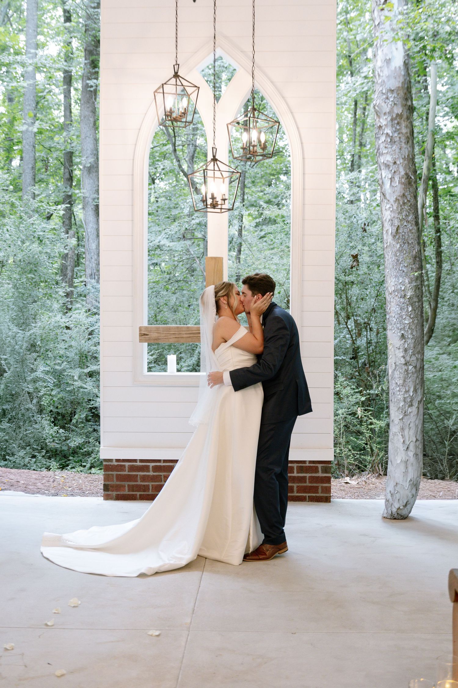 Bride and groom share first kiss at outdoor wedding chapel surrounded by lush green trees with hanging lanterns.