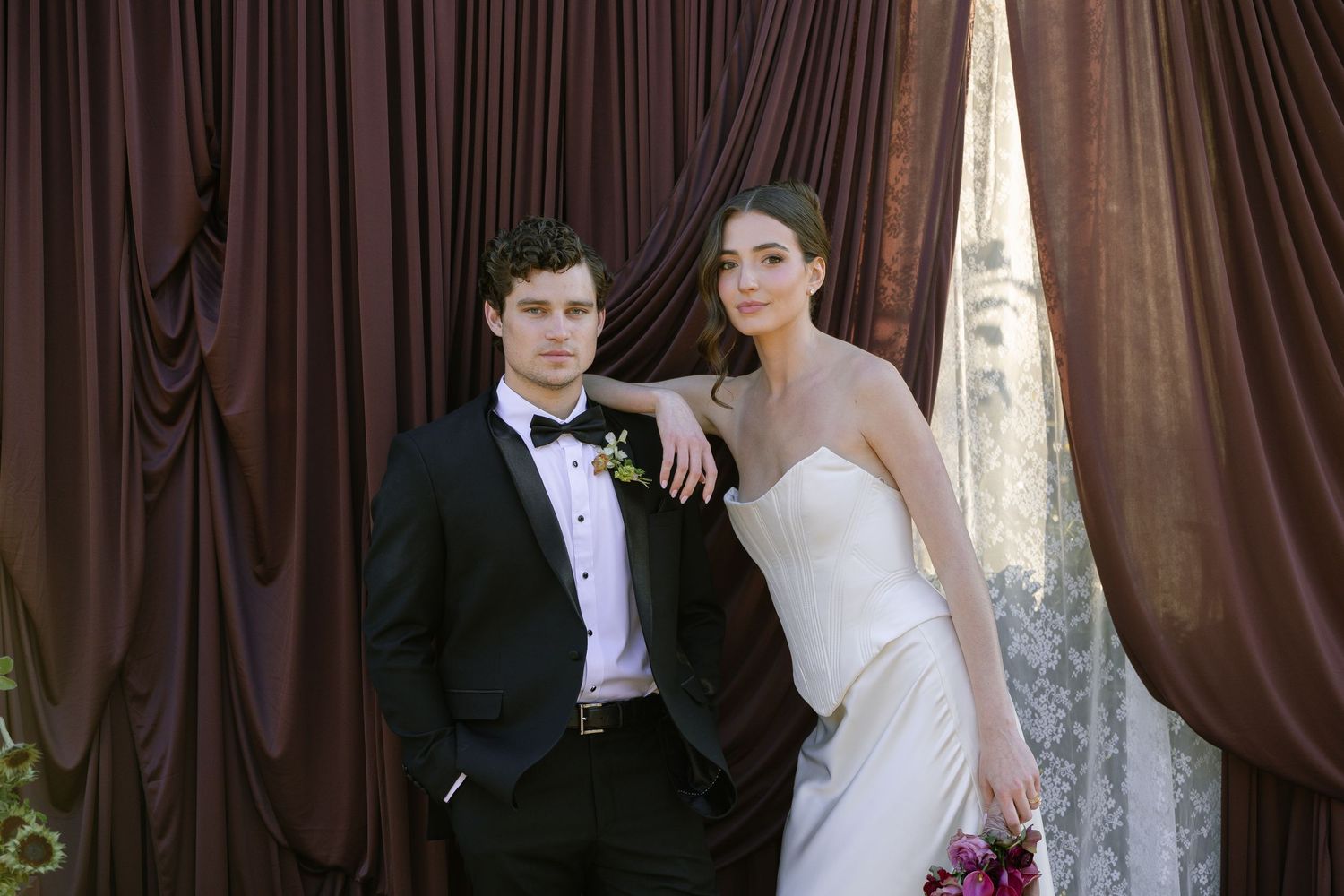Elegant bride and groom posing in front of luxurious dark brown draped curtains at a formal wedding ceremony.