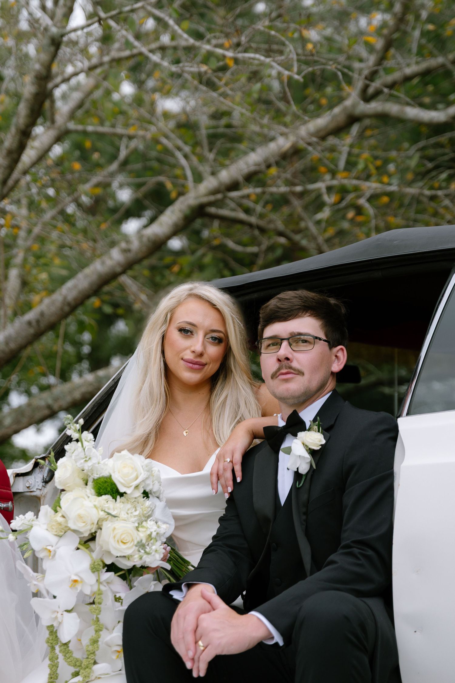 Bride and groom sitting together by a white limo, she holds white roses bouquet, he wears a black tuxedo with bow tie.