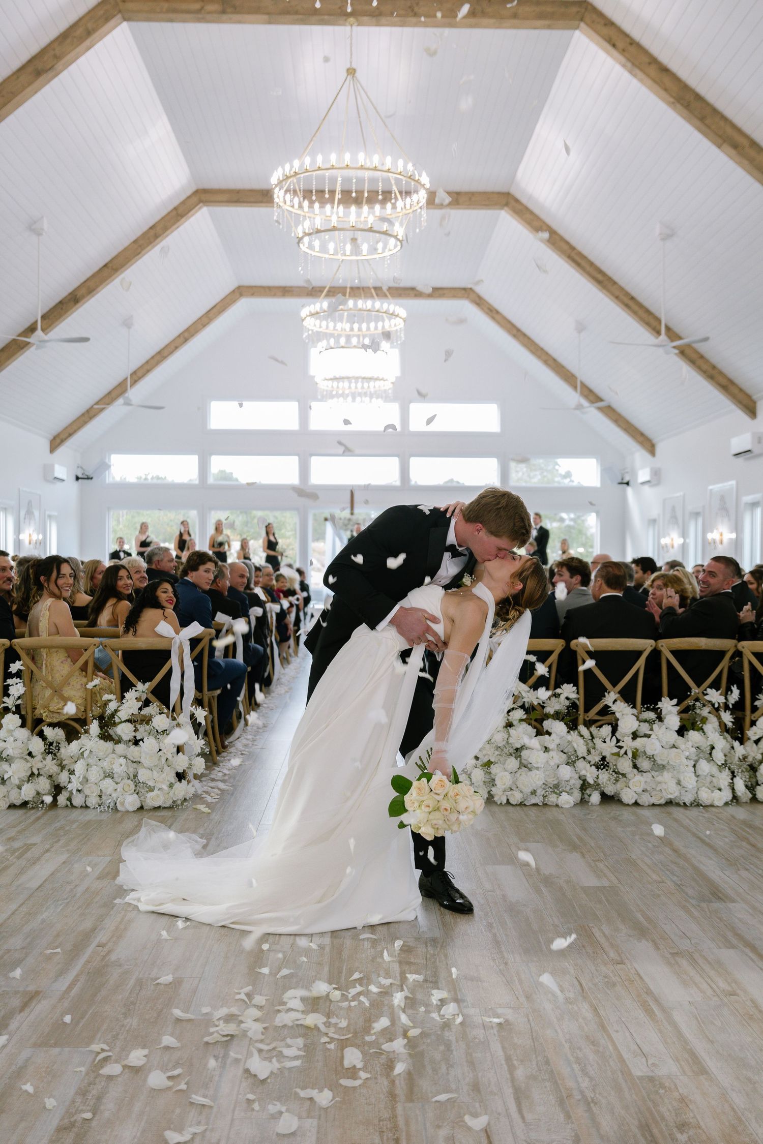 Newlyweds share a dip kiss at altar with white rose aisle decor and chandelier in chapel.