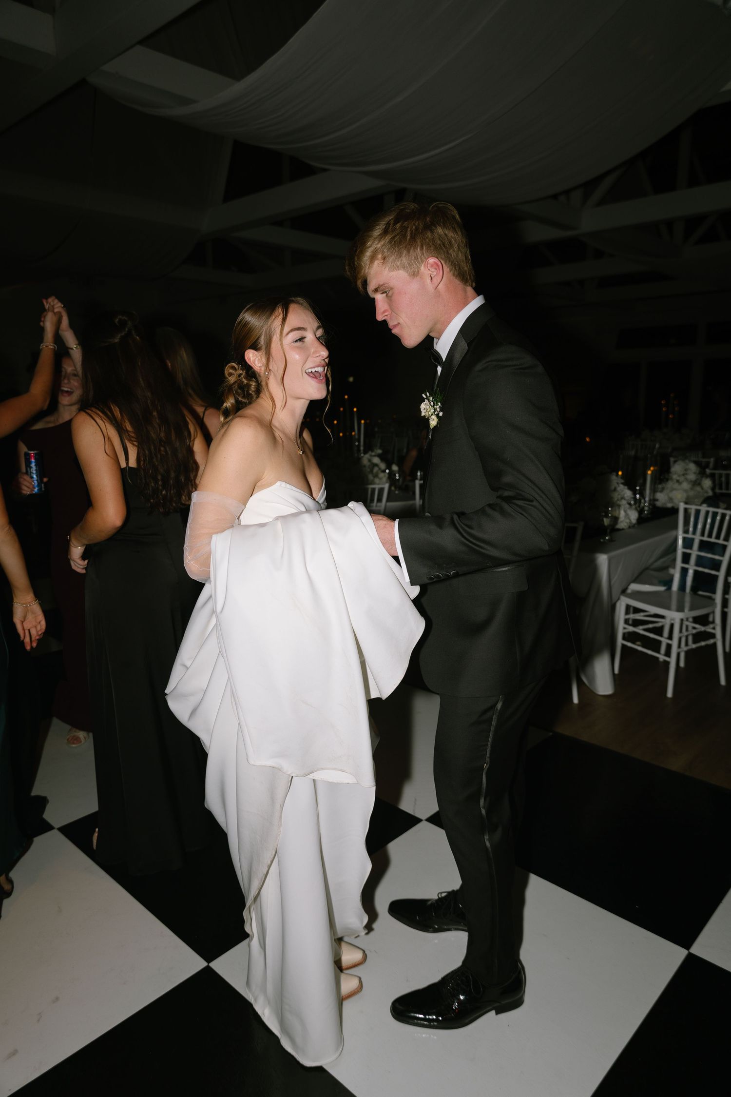 Bride and groom dancing joyfully on checkered black and white dance floor at wedding reception.