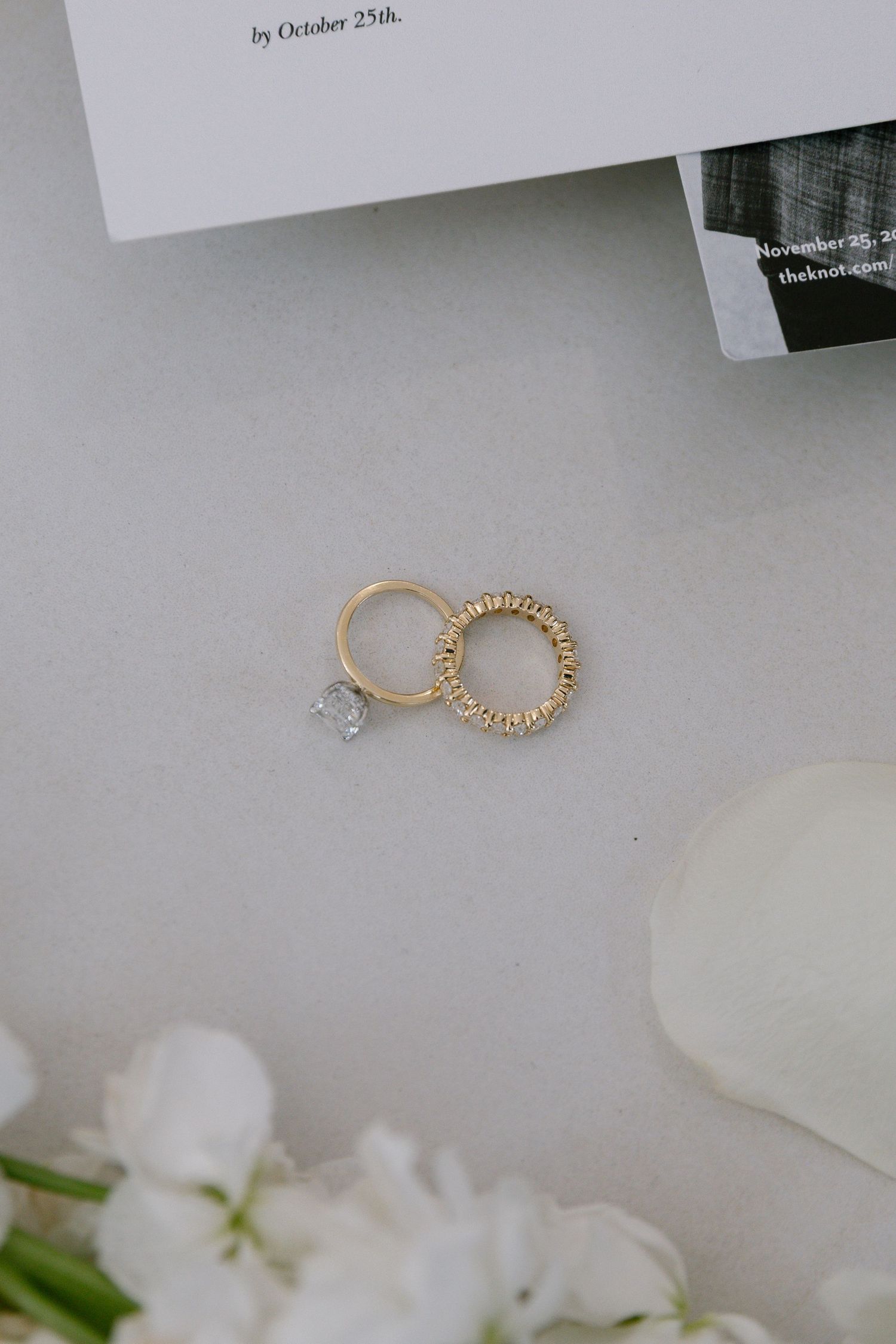 Gold wedding bands and diamond engagement ring on gray surface with white flowers and wedding stationery.