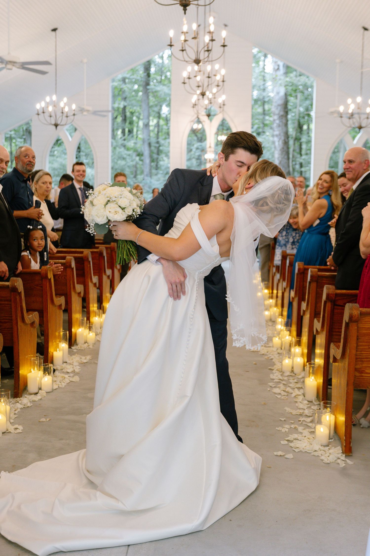 Bride and groom sharing a romantic dip kiss at the altar of a candlelit white chapel.