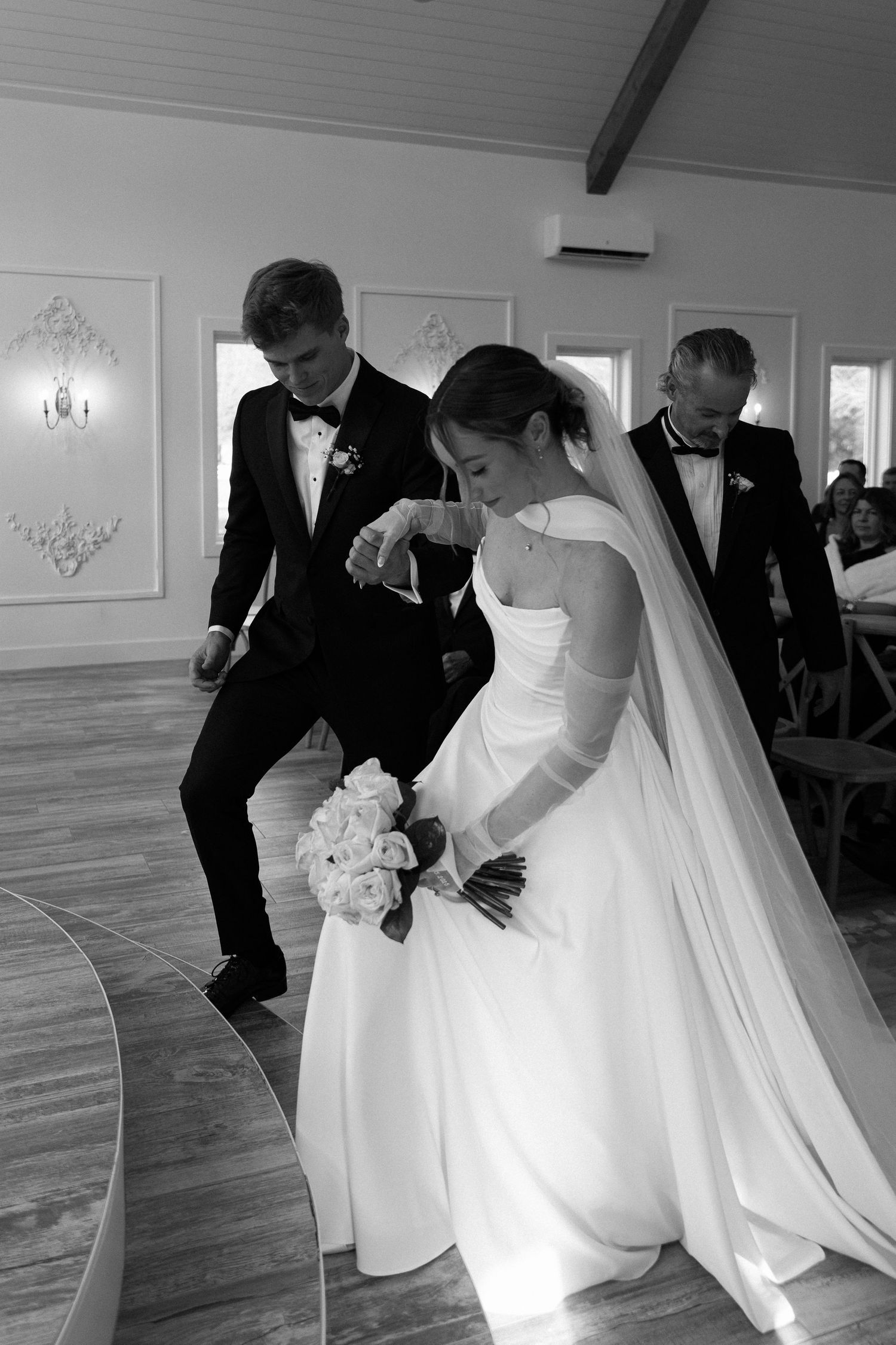 Bride and groom kneeling together during wedding ceremony in black and white photo with bouquet.