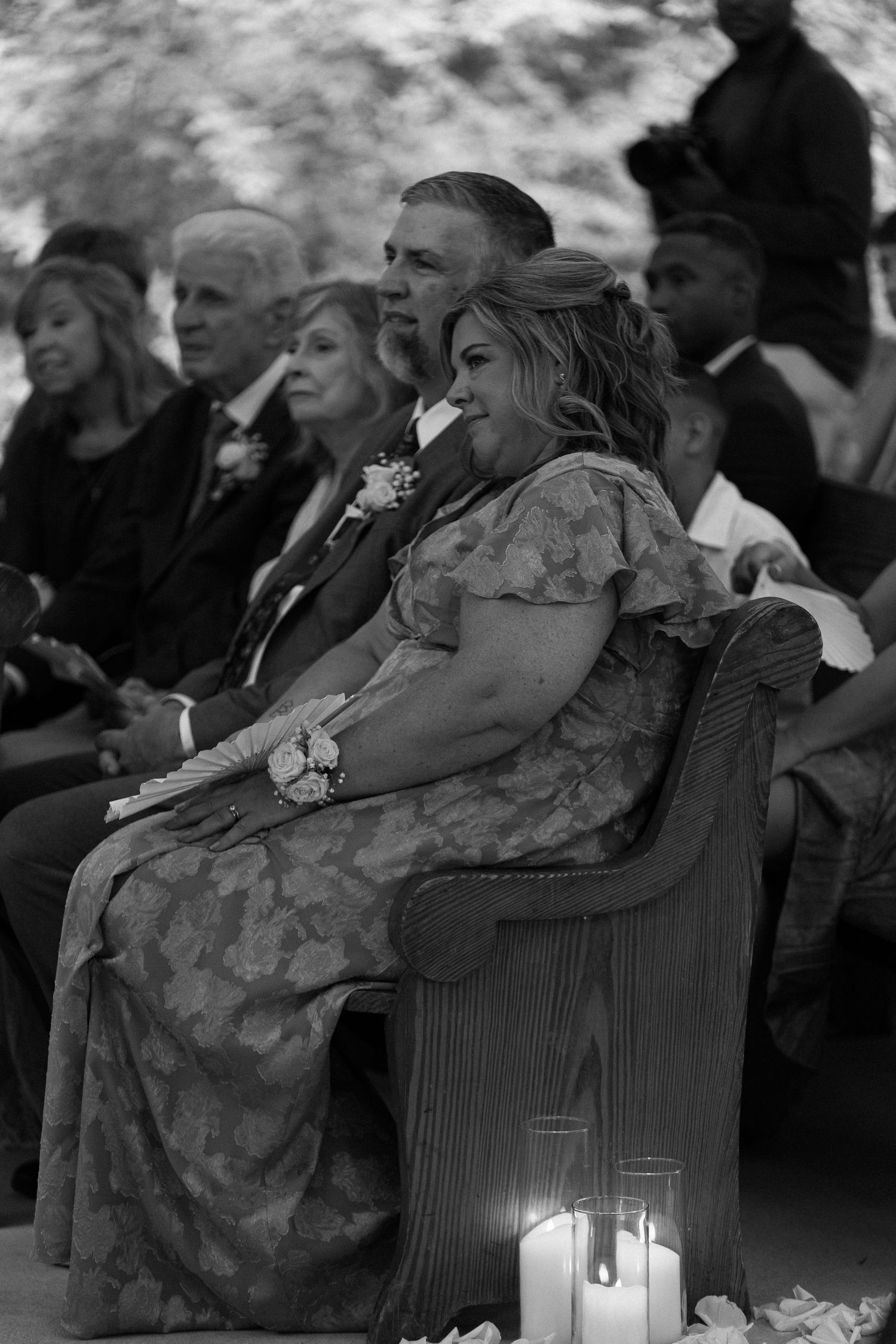 Black and white photo of wedding guests seated on wooden pews, with lit candles in foreground during outdoor ceremony.