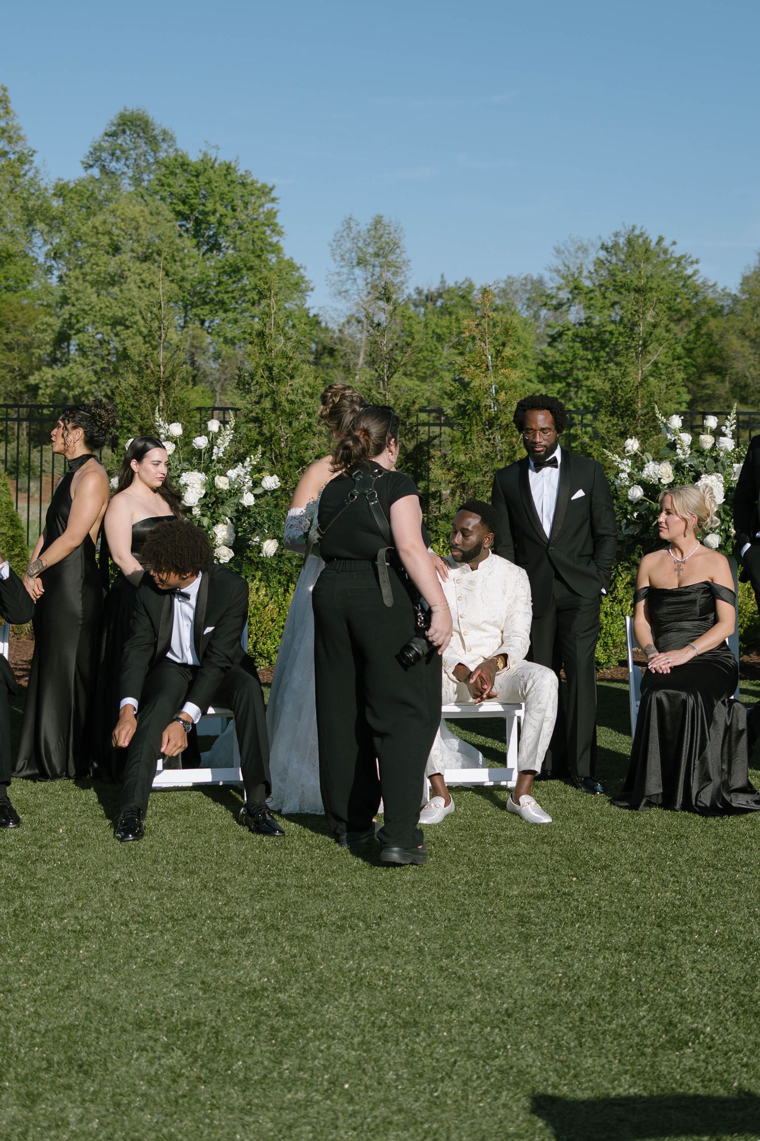 Outdoor wedding ceremony with guests in black formal attire on green lawn, groom seated in white suit near floral arch.