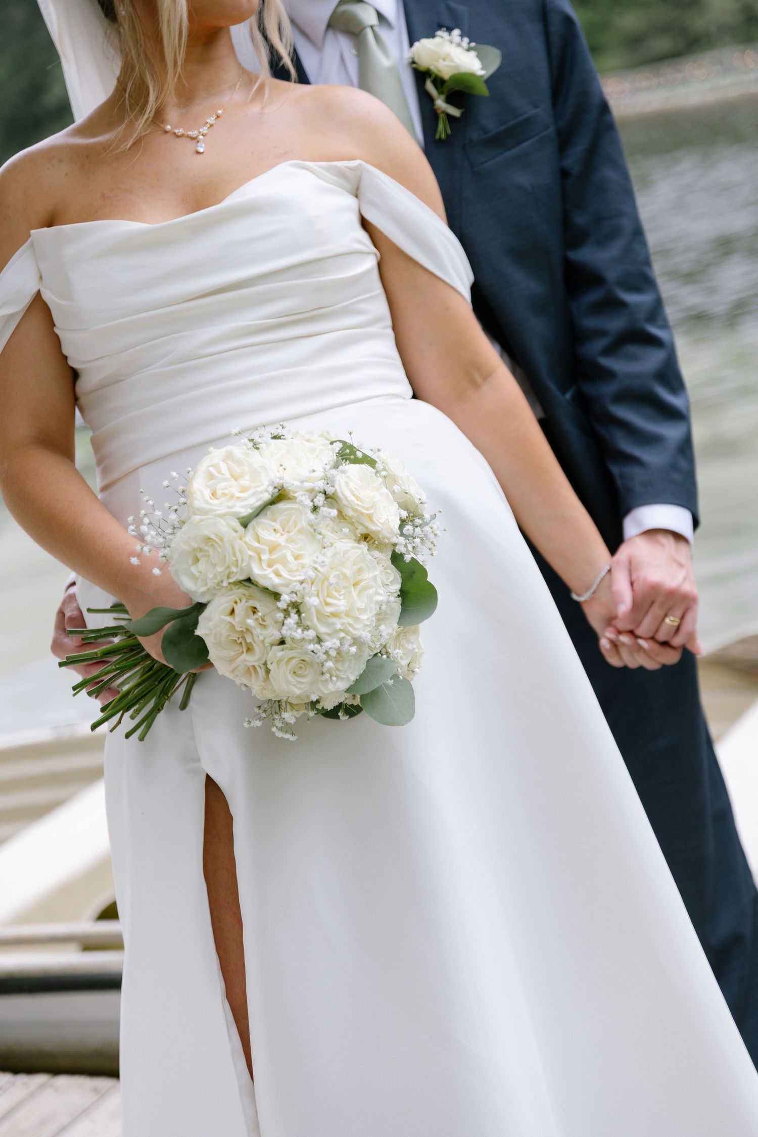 Bride holding white rose bouquet in off-shoulder wedding dress holding hands with groom in navy suit by waterfront.