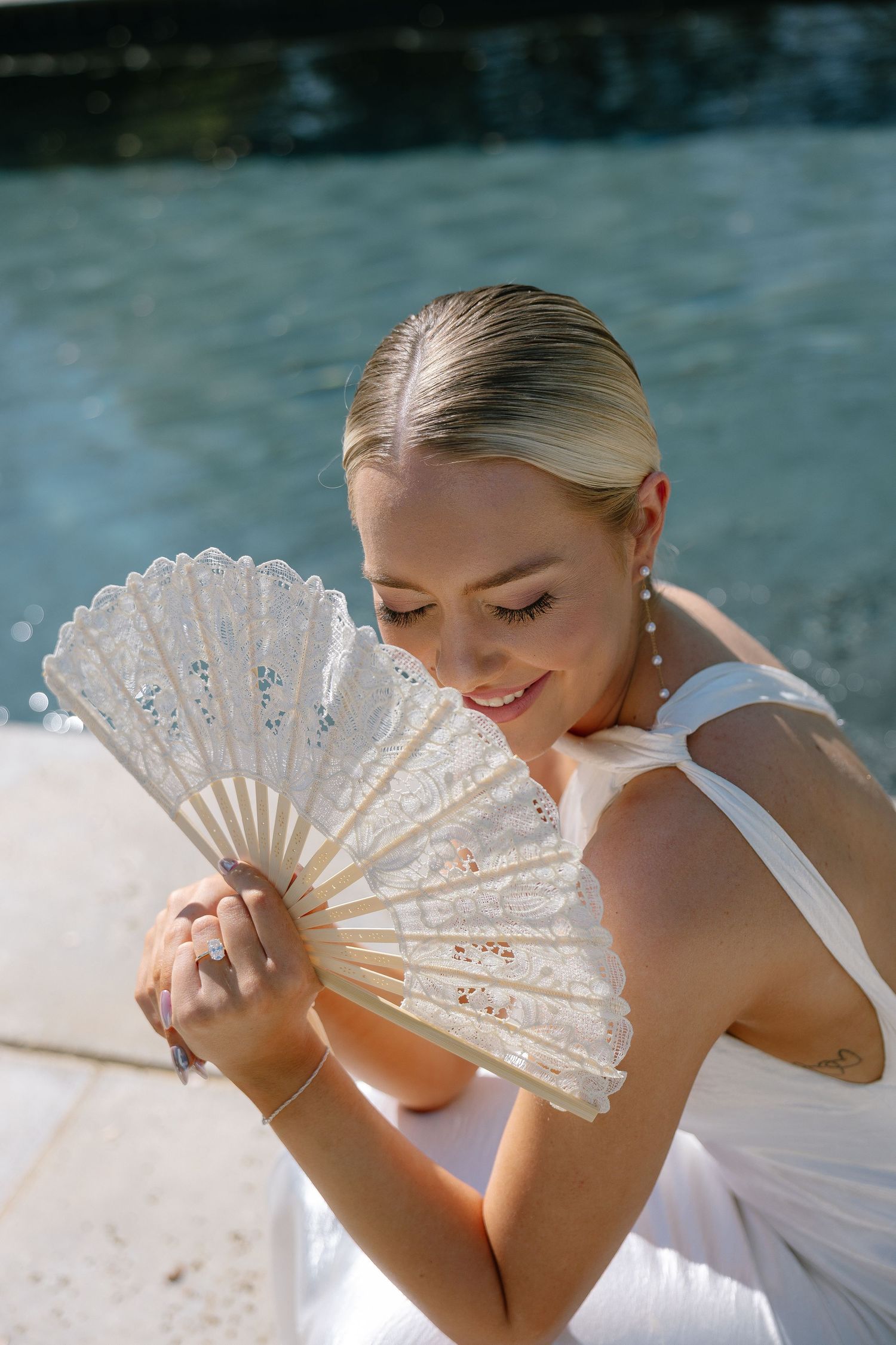 Elegant blonde woman in white dress holding a decorative lace hand fan near a pool, smiling softly.