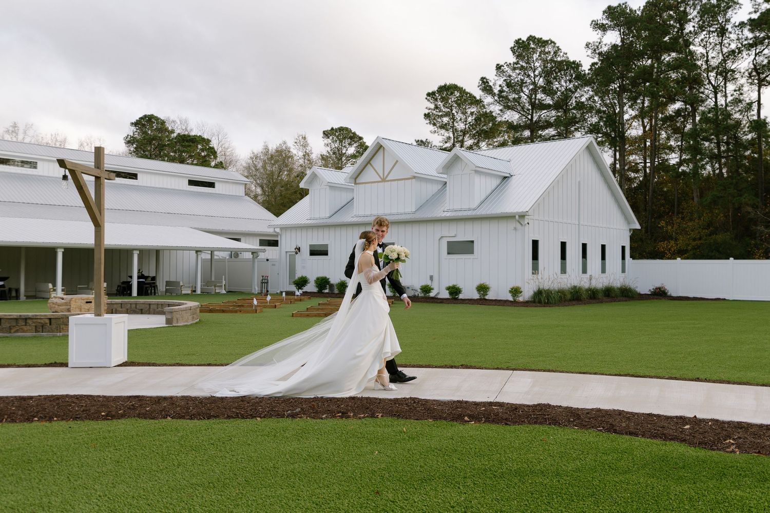 Bride and groom walk hand in hand outside a elegant white barn wedding venue on a cloudy day.