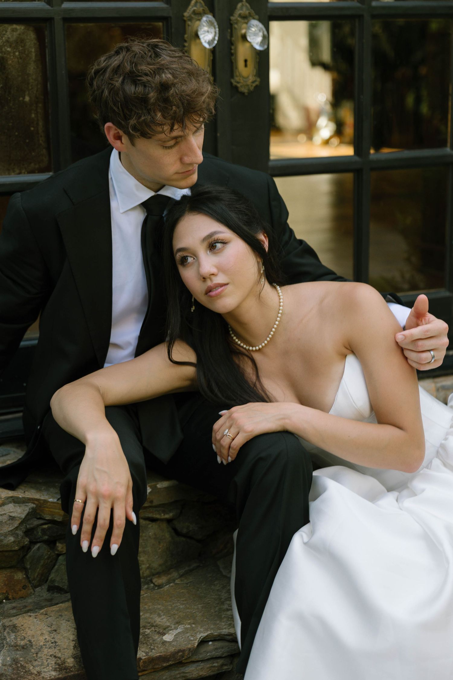 A bride in white gown and pearl necklace sits with groom in black suit on stone steps in an intimate wedding portrait.