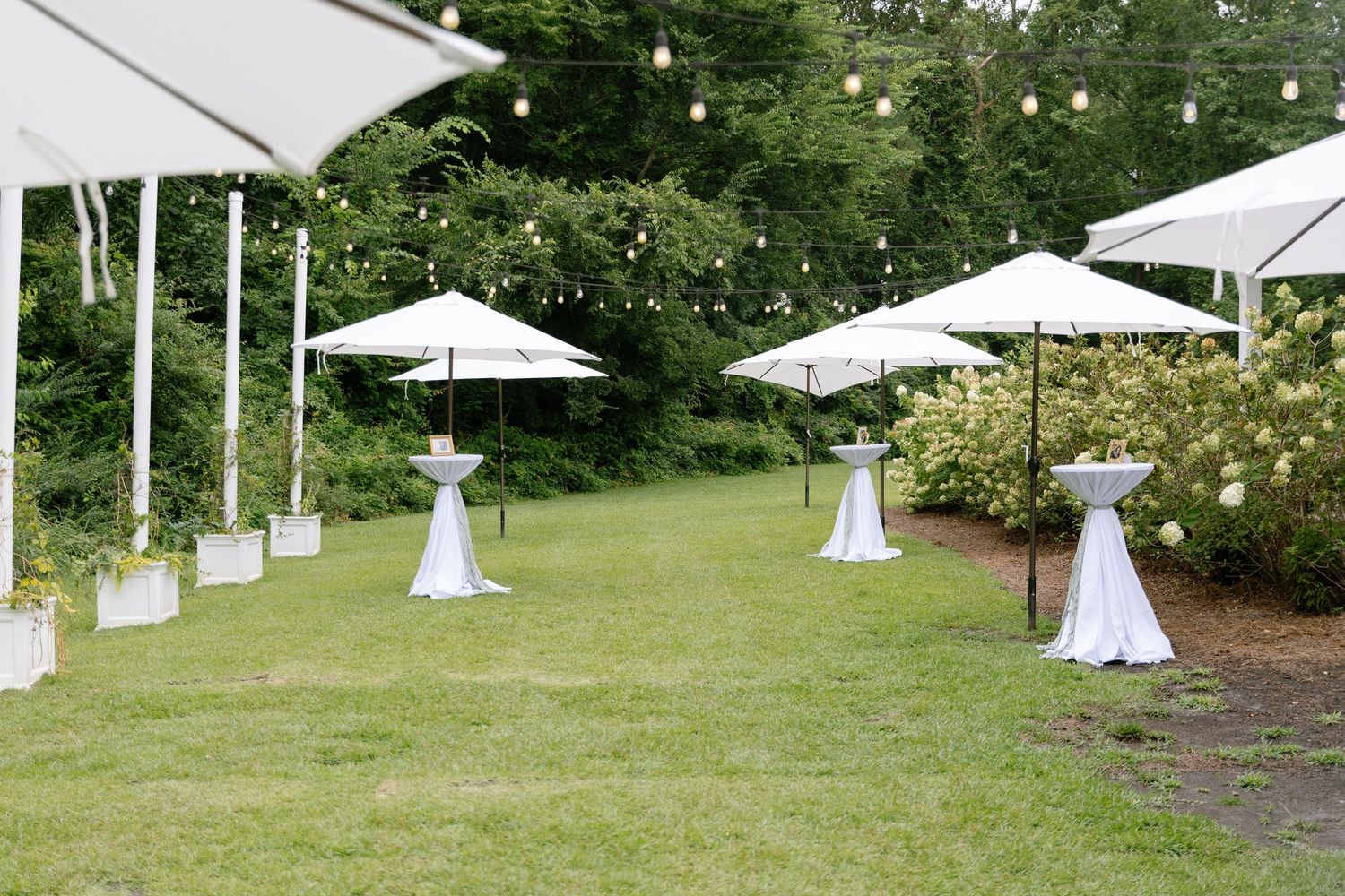 Outdoor wedding cocktail hour setup with white umbrellas, draped high-top tables, string lights, and lush green garden backdrop.