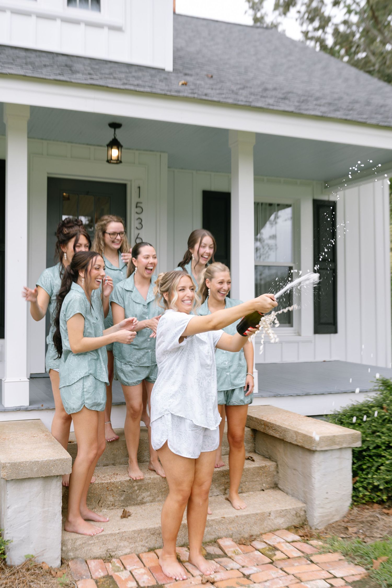 Bride popping champagne with bridesmaids in matching sage green pajamas on a white farmhouse porch.