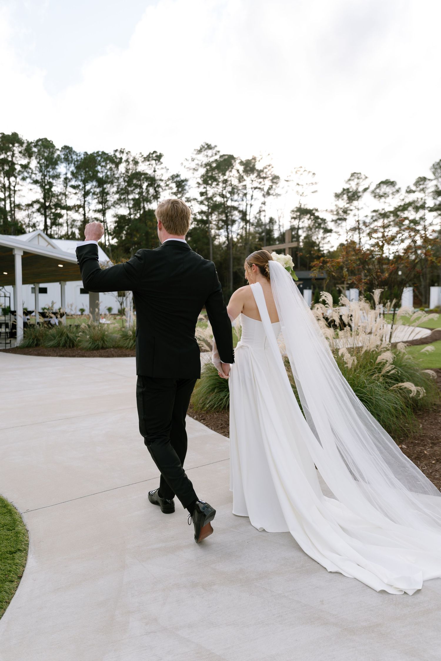 Bride and groom walk hand-in-hand on pathway, bride wearing long cathedral veil trailing behind her.