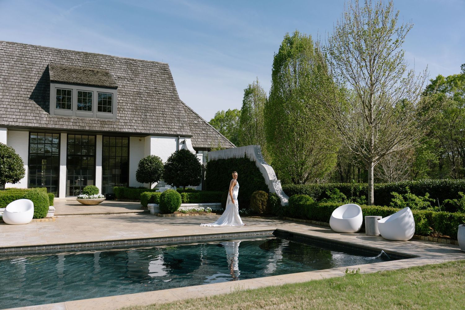 Bride in white gown standing by a luxury outdoor pool beside an elegant Hamptons-style home with manicured hedges.