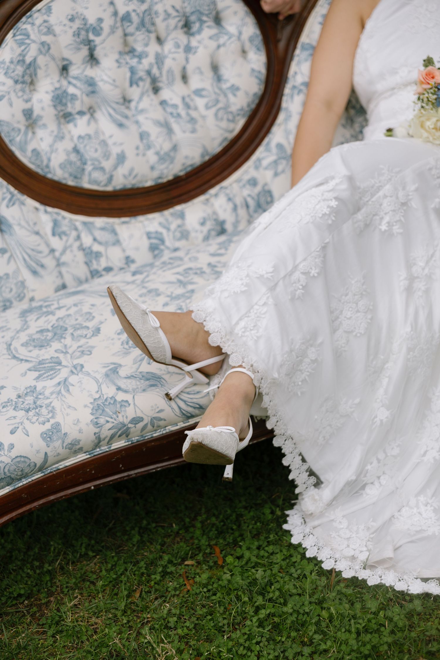 Bride in white lace gown seated on vintage floral chaise lounge, wearing elegant white heels on green grass.