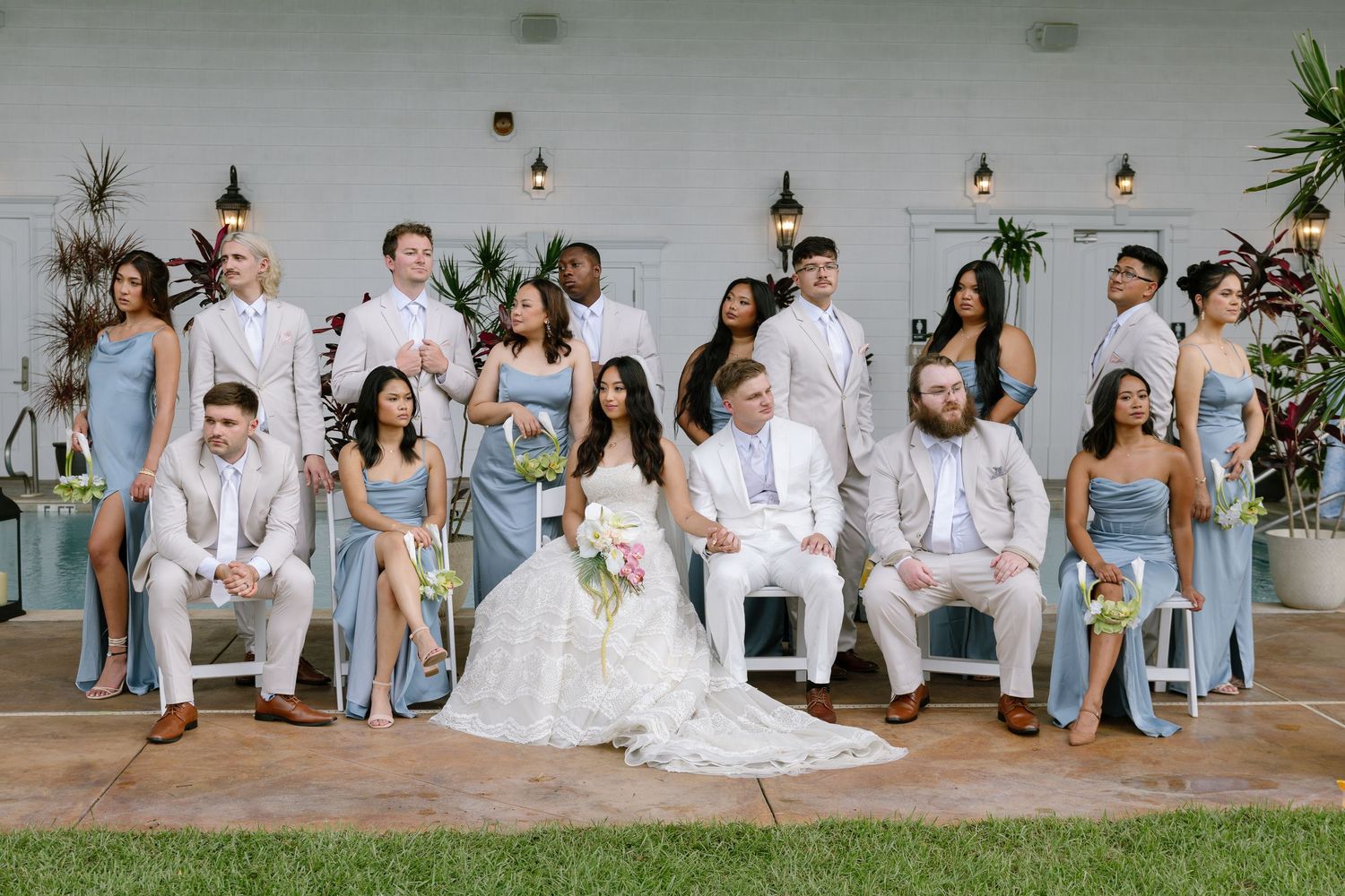 Wedding party poses in light blue and white attire, bride and groom seated center, outdoors at elegant venue.