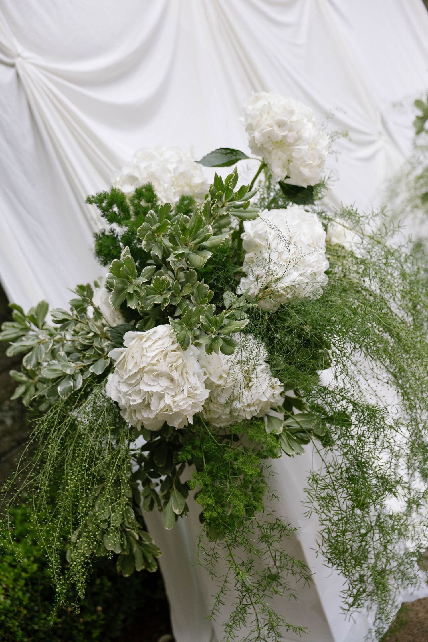 Elegant white floral arrangement with hydrangeas and lush greenery against draped white fabric backdrop.