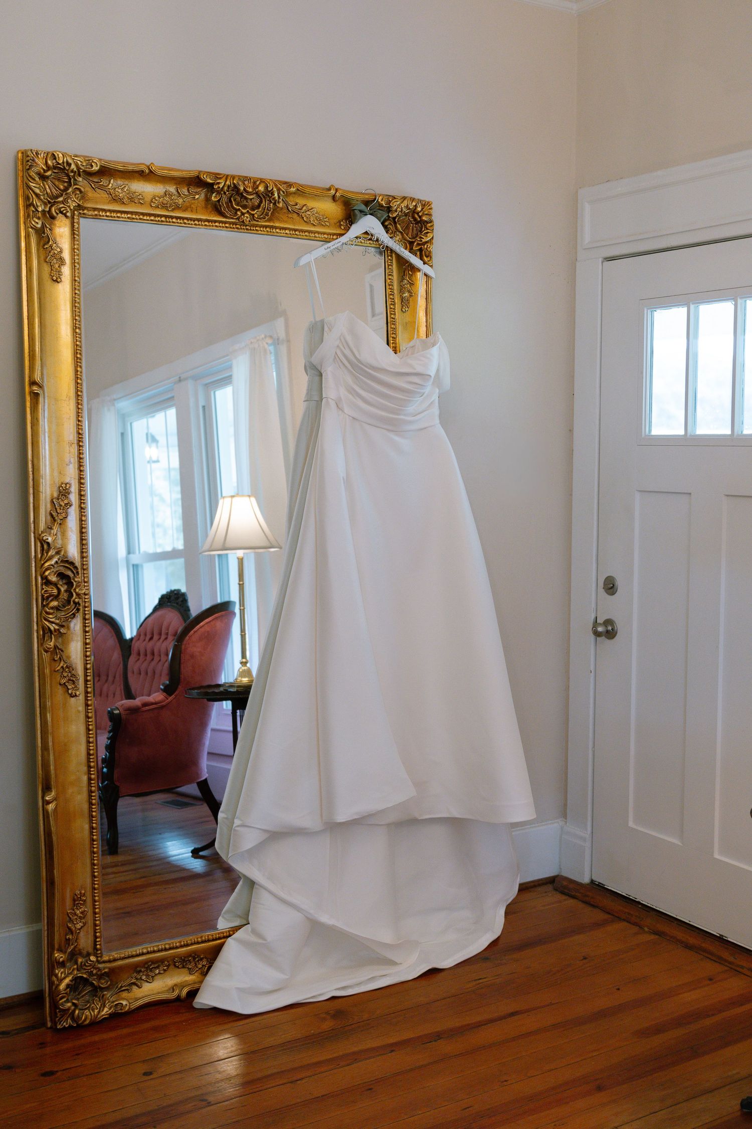 White wedding gown hanging on a gold ornate mirror in a bright room with hardwood floors.