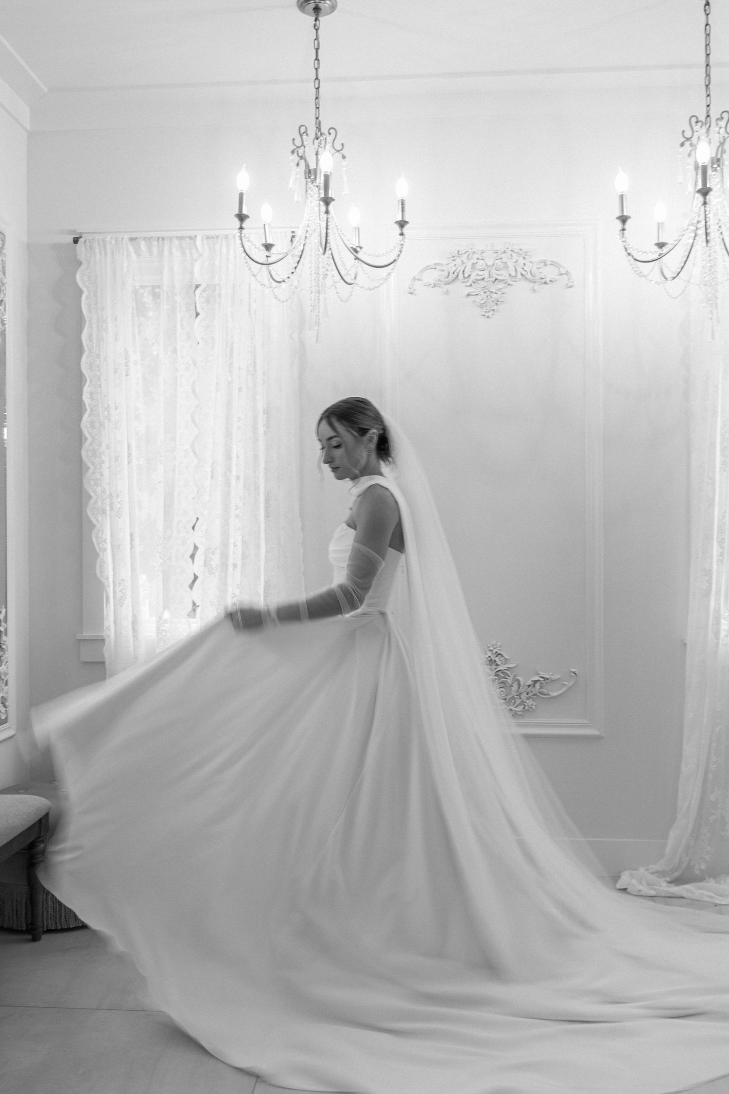 Bride twirling in a flowing white wedding gown with cathedral veil in an elegant chandelier-lit room.