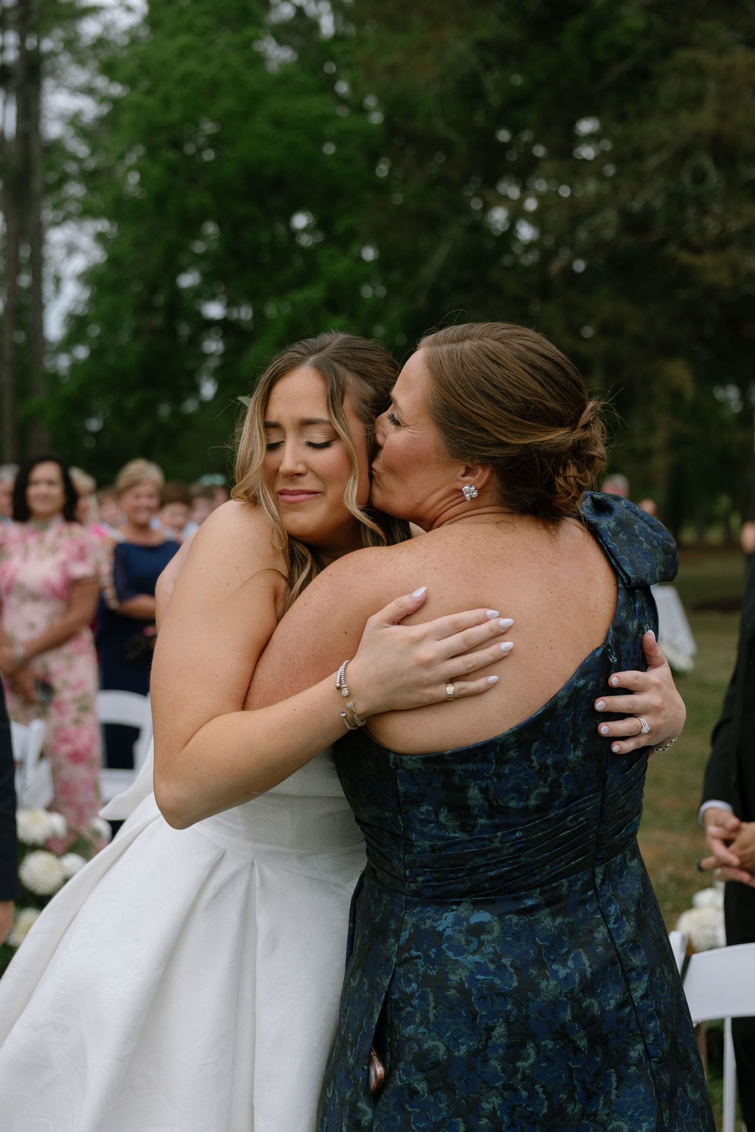 Bride in white gown embracing a woman in navy floral dress at outdoor wedding ceremony with guests in background.