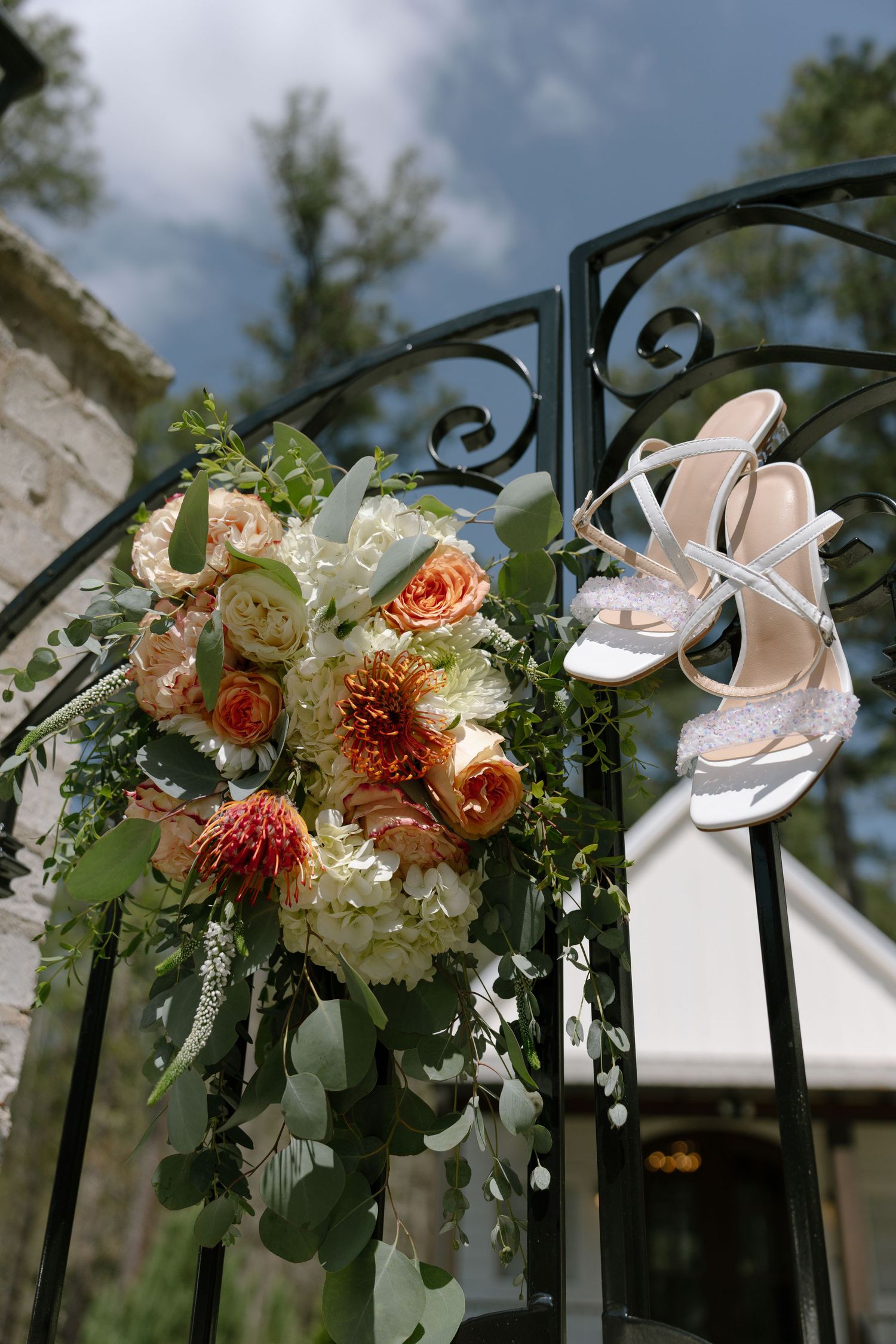 Bridal bouquet with peach roses and white hydrangeas hanging on ornate black iron gate beside elegant white strappy wedding heels.