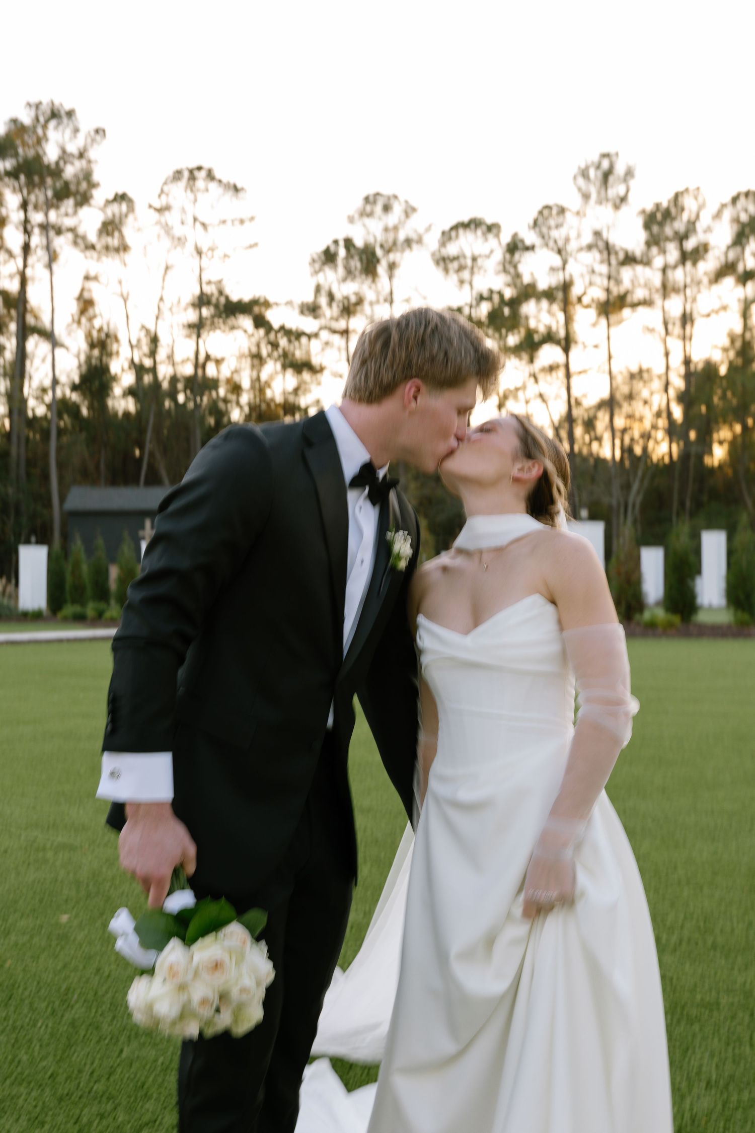 Newlyweds share a kiss on a green lawn at sunset, groom holding white bridal bouquet.