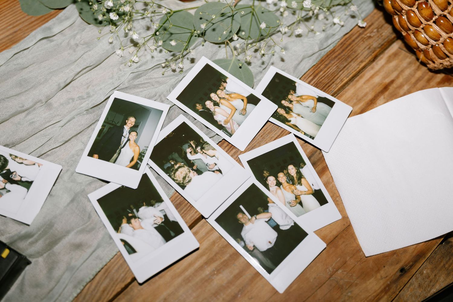 Scattered Polaroid photos of wedding couples displayed on rustic wooden table with eucalyptus decor.
