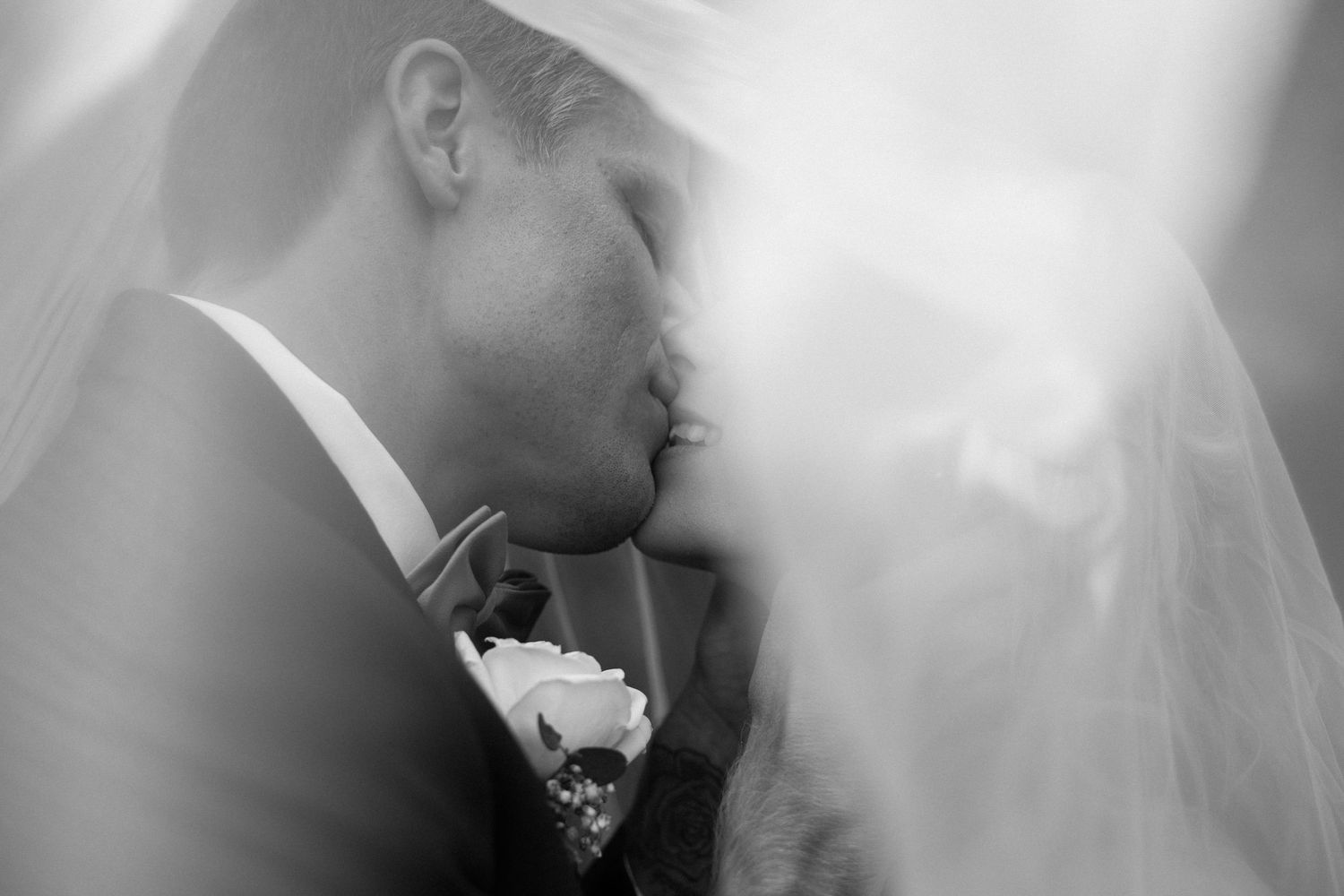 Black and white photo of a bride and groom sharing a romantic kiss beneath a flowing wedding veil.