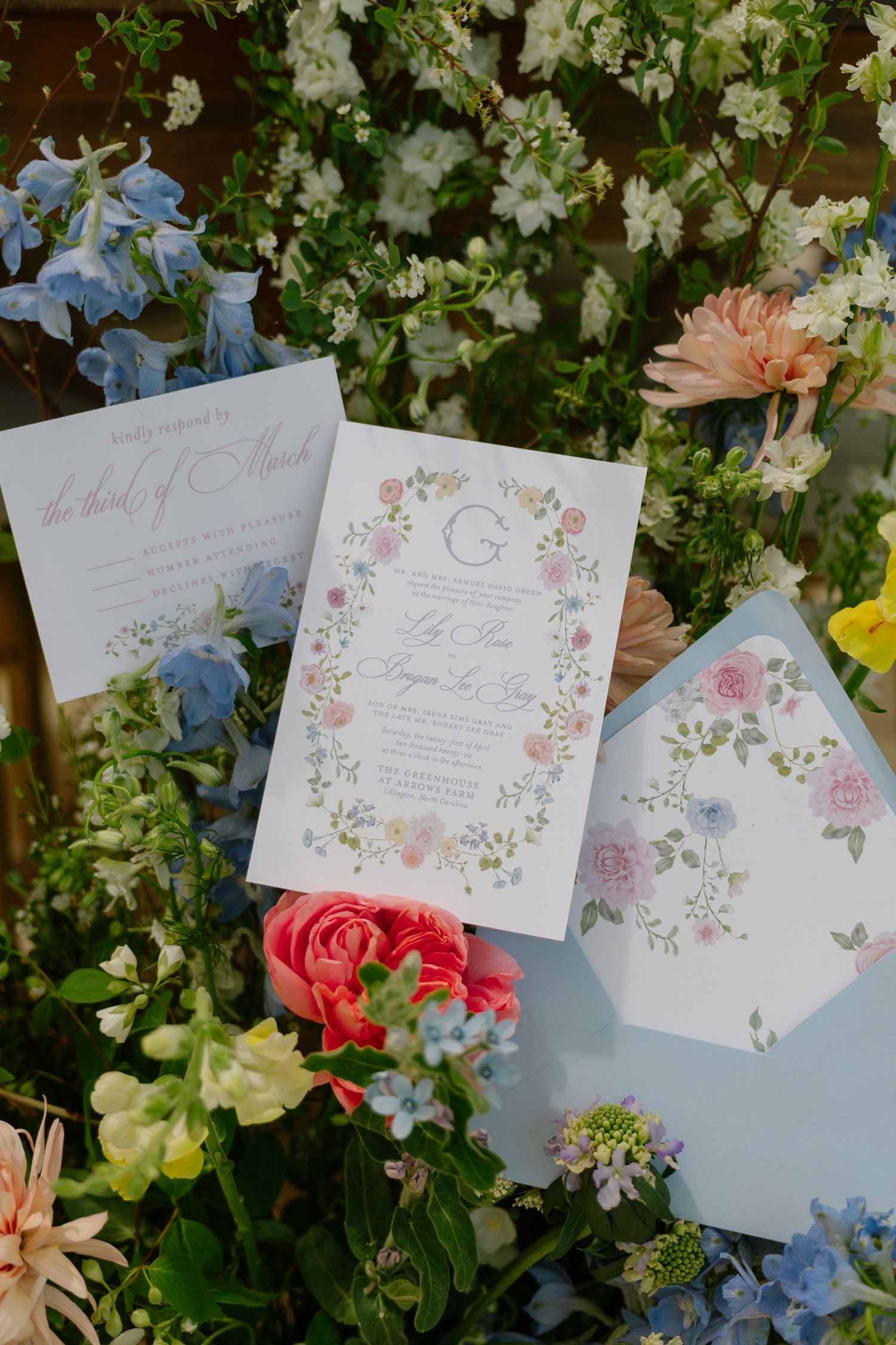 Floral wedding invitation suite with pastel pink, blue flowers and monogram G, displayed among fresh garden blooms.