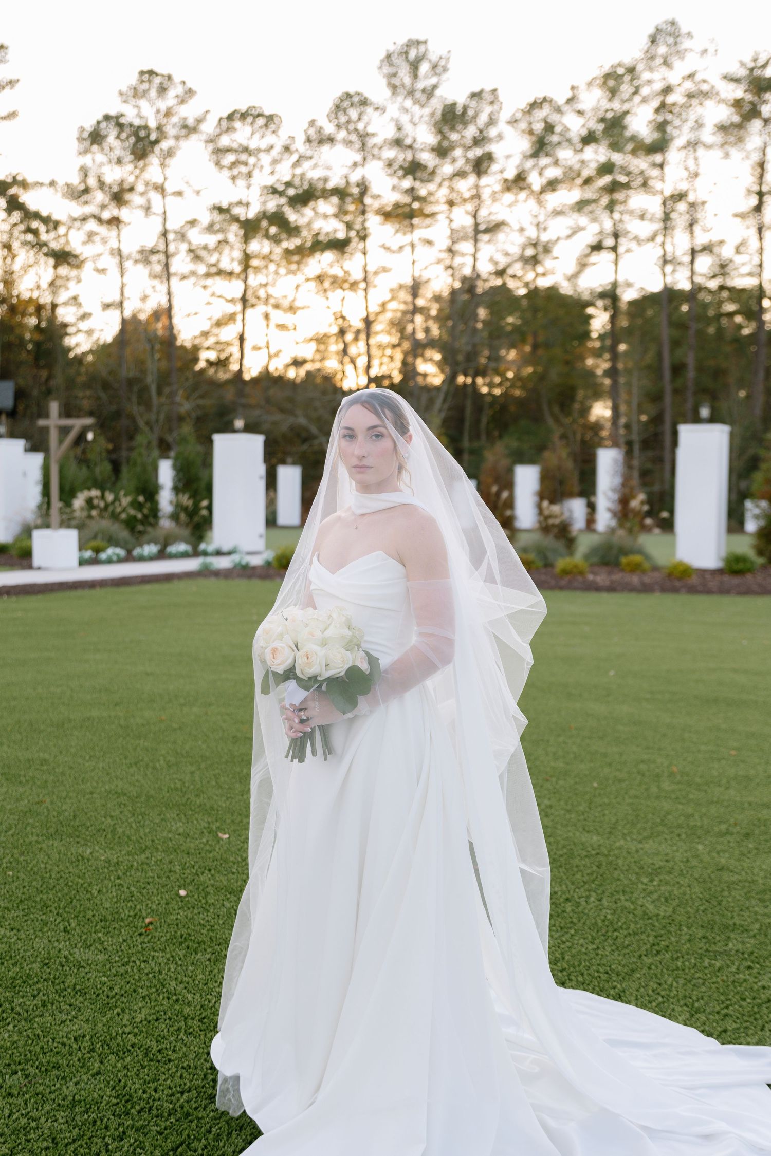 Bride in strapless white gown and long veil holds bouquet on green lawn at sunset with white pillars behind.
