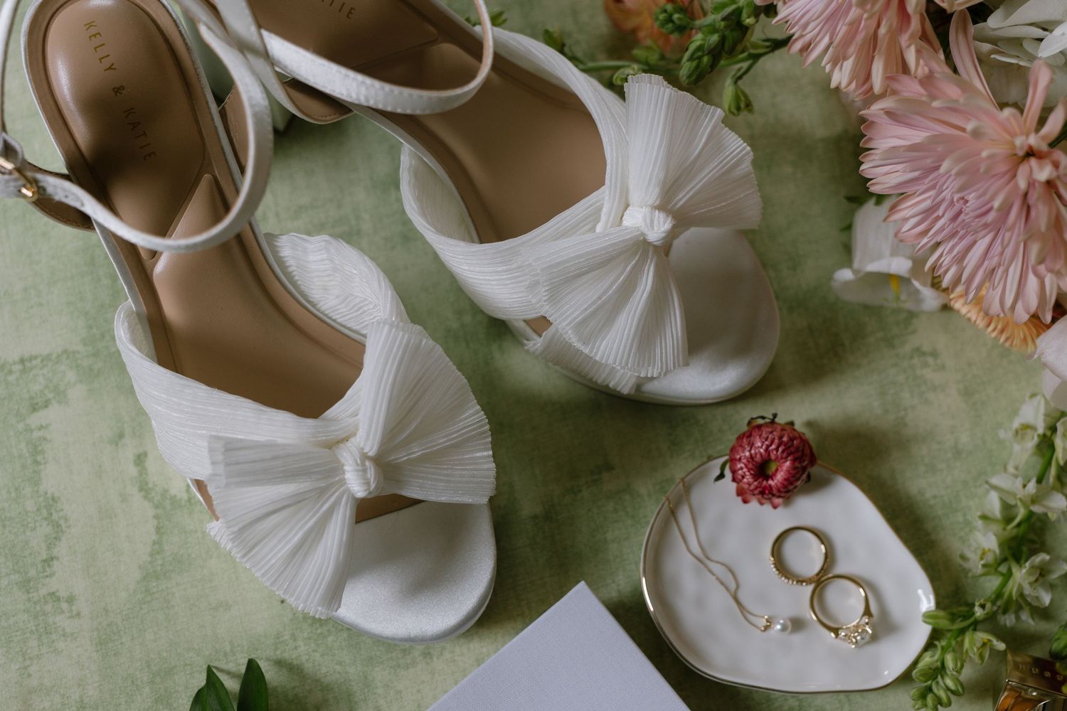 White bridal heels with bow detail beside gold rings and pink flowers on a green surface.