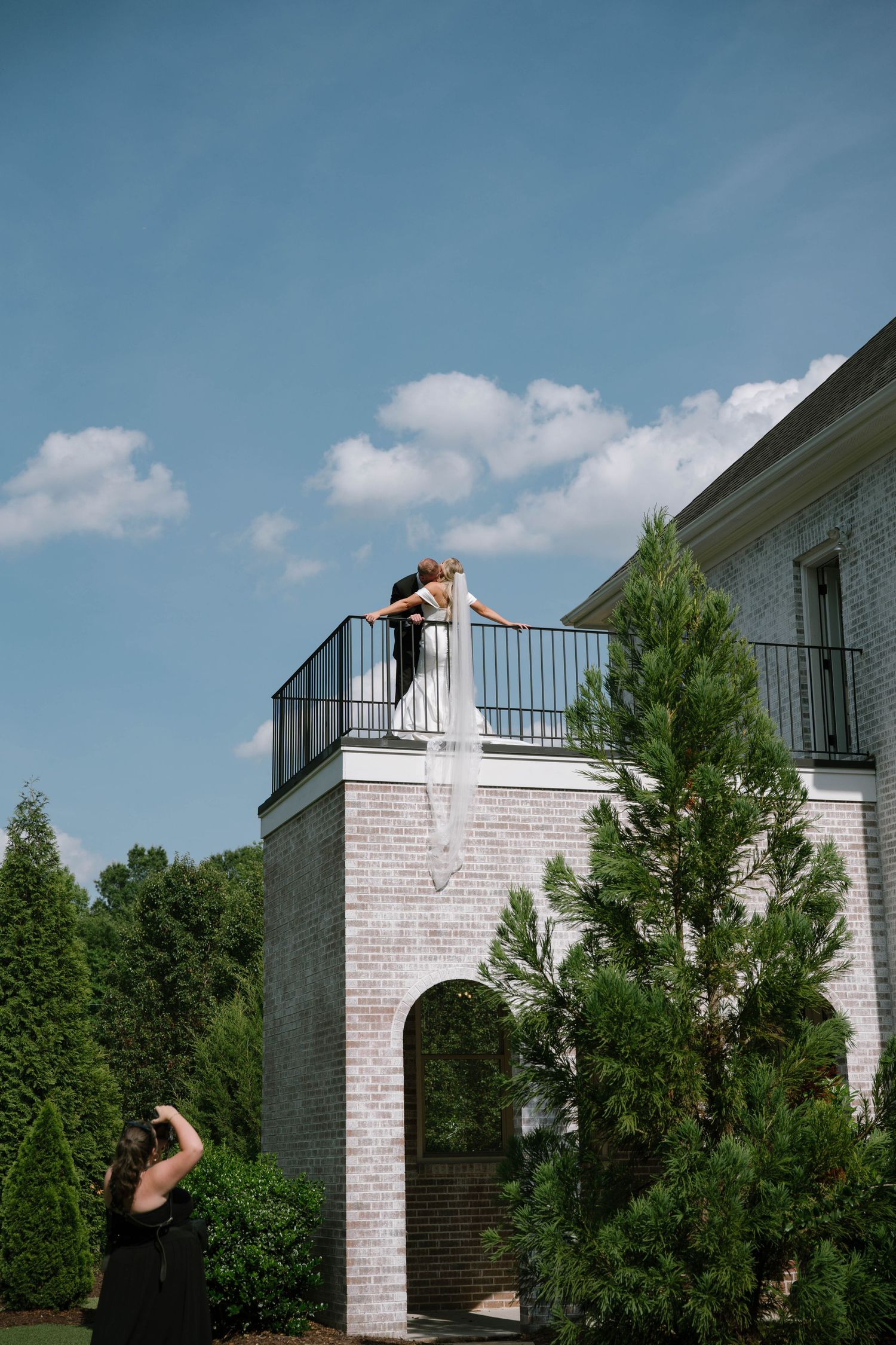 Bride and groom embrace on a balcony of a brick building while a photographer captures the moment below.