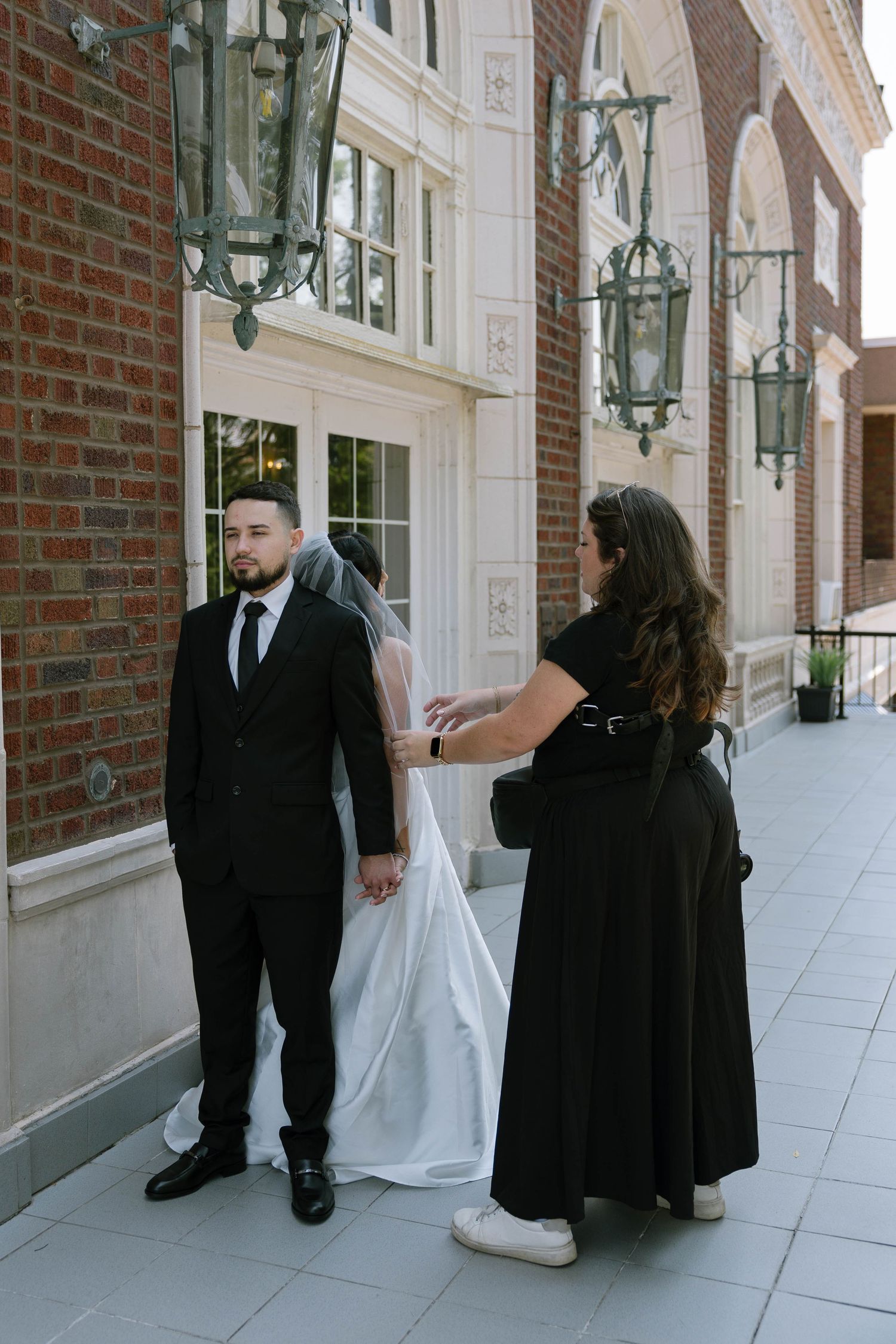 A wedding coordinator adjusts a groom's veil outside a brick building with ornate lanterns.