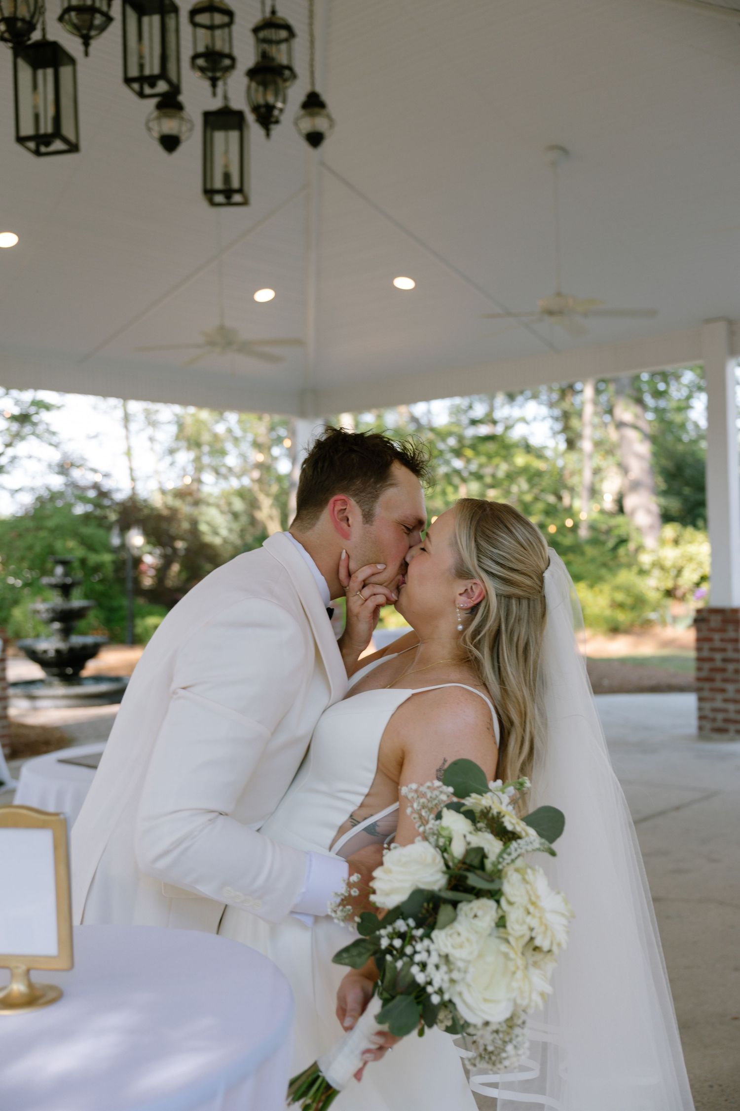 Bride and groom share first kiss under elegant pavilion, bride holding white rose bouquet with eucalyptus greenery.