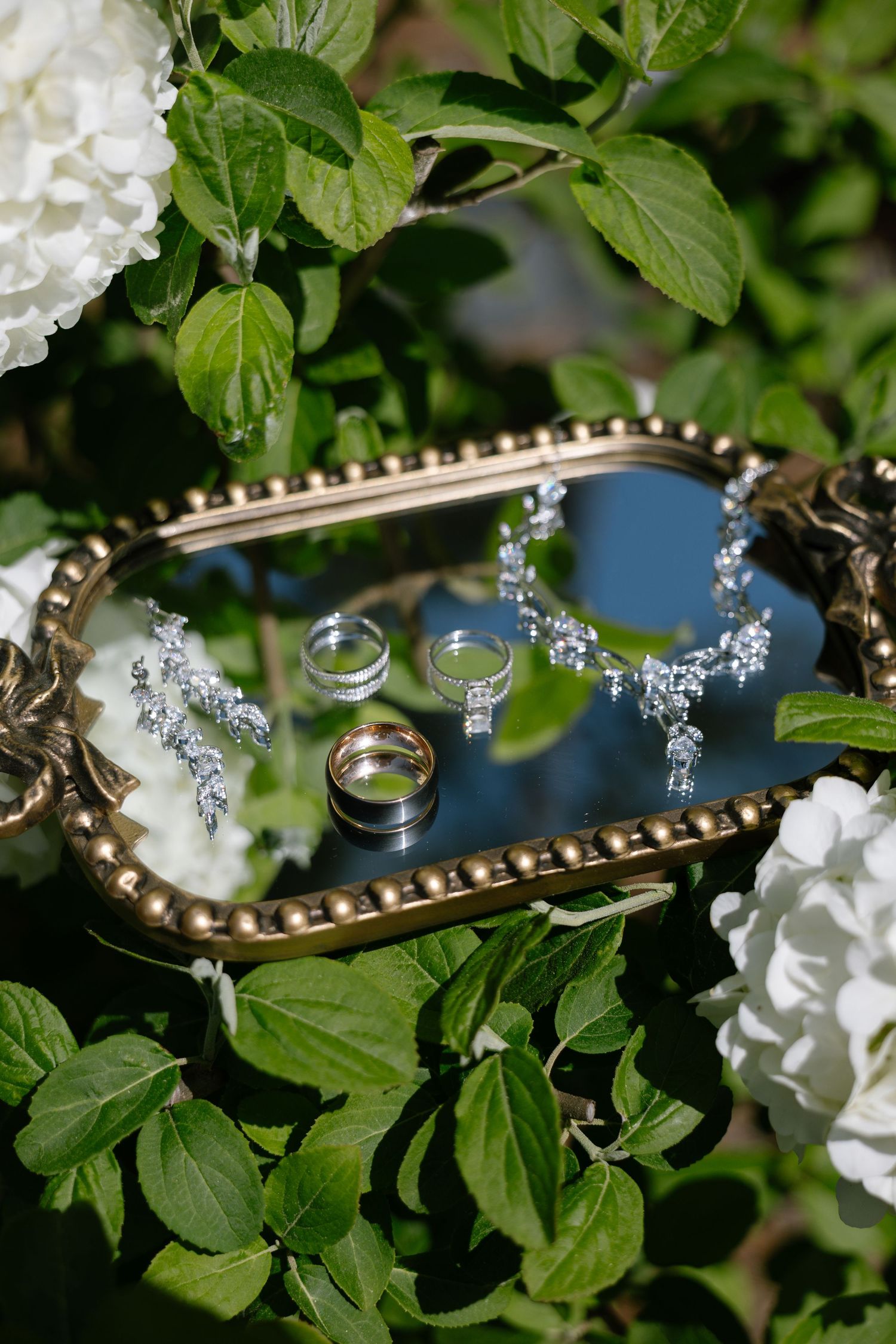 Wedding jewelry displayed on a gold mirrored tray with diamond necklace, earrings, and rings surrounded by white hydrangeas.
