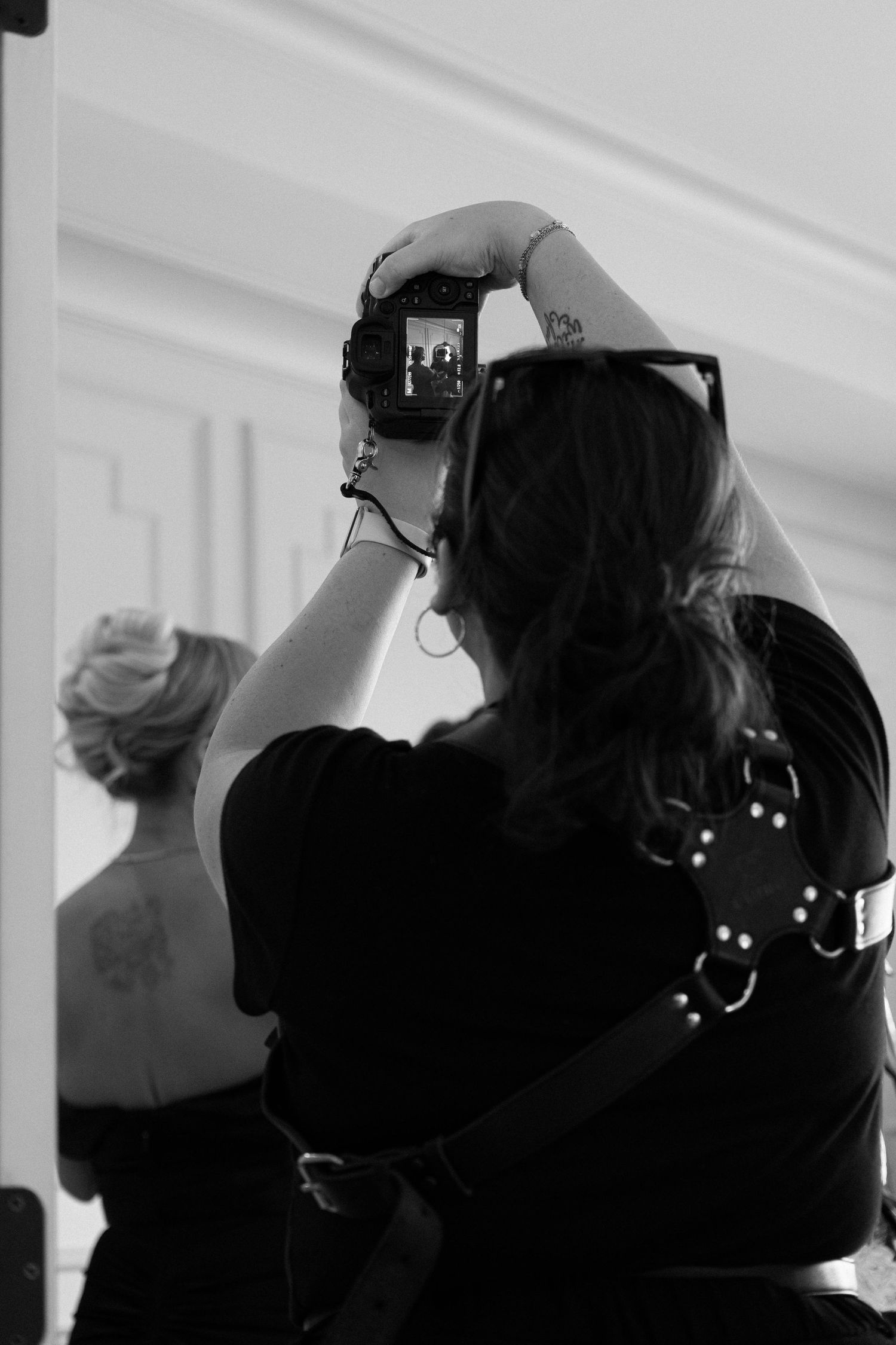 Black and white photo of a woman photographing with a camera held overhead, another woman with updo visible in background.