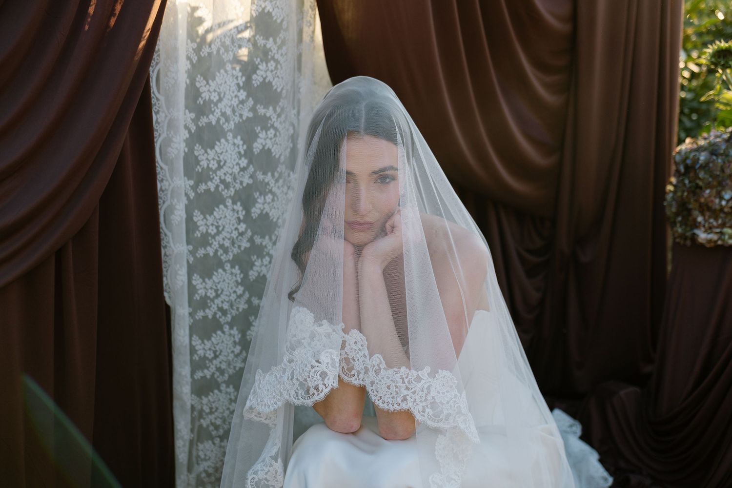 Bride in lace veil and white wedding gown poses pensively beside elegant brown drapes in soft natural light.