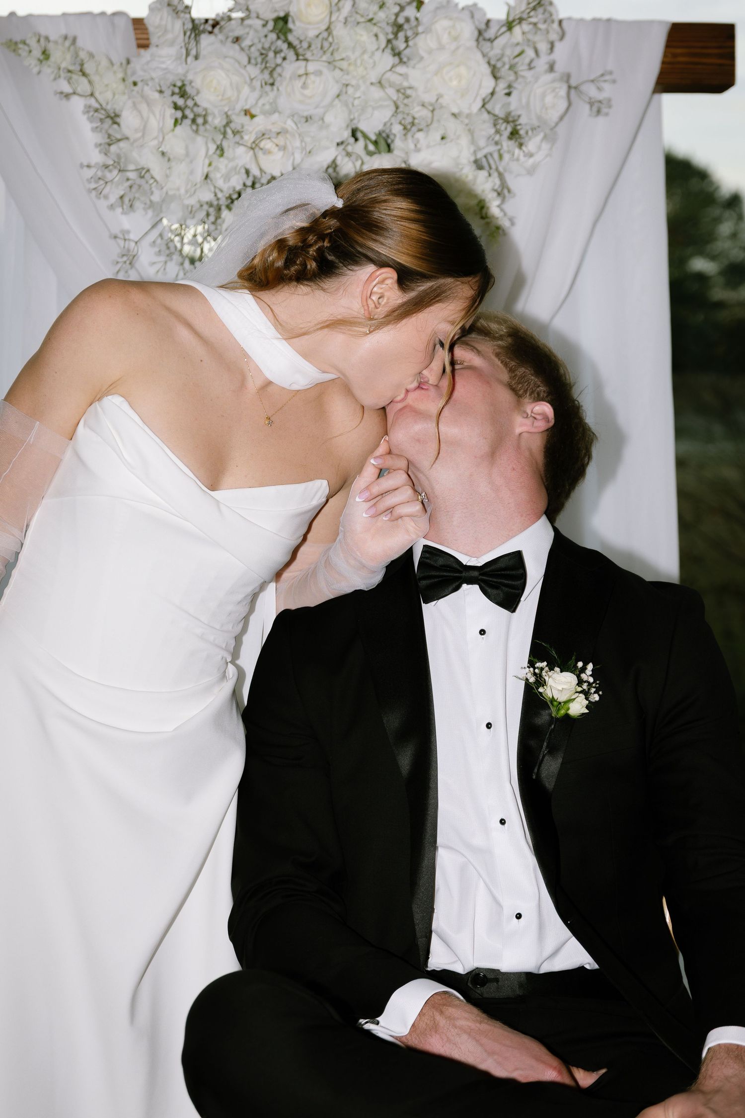Bride in white gown kissing groom in black tuxedo at wedding altar with white floral backdrop.