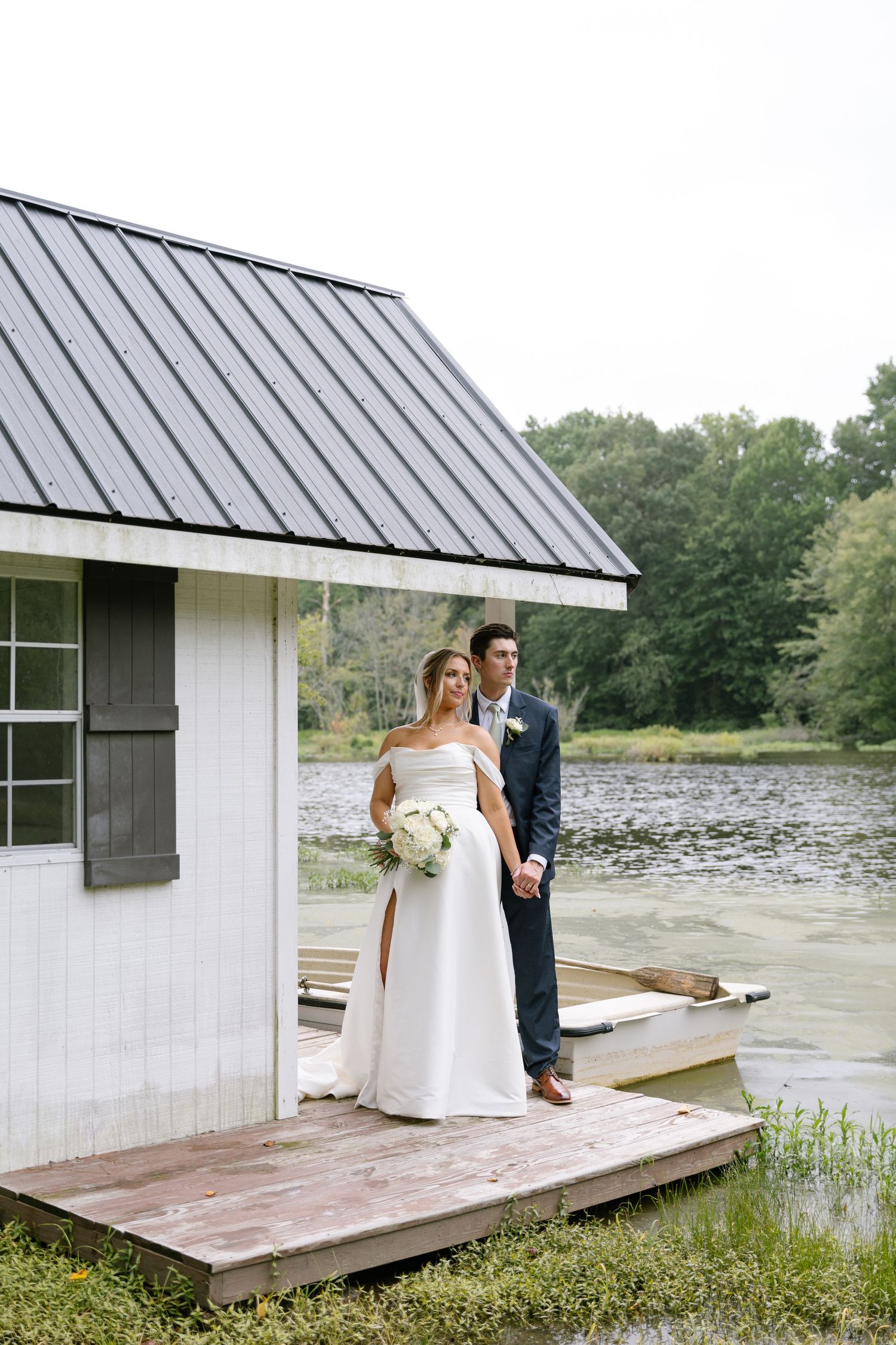 Bride and groom posing on a dock beside a white boathouse with a serene lake backdrop.