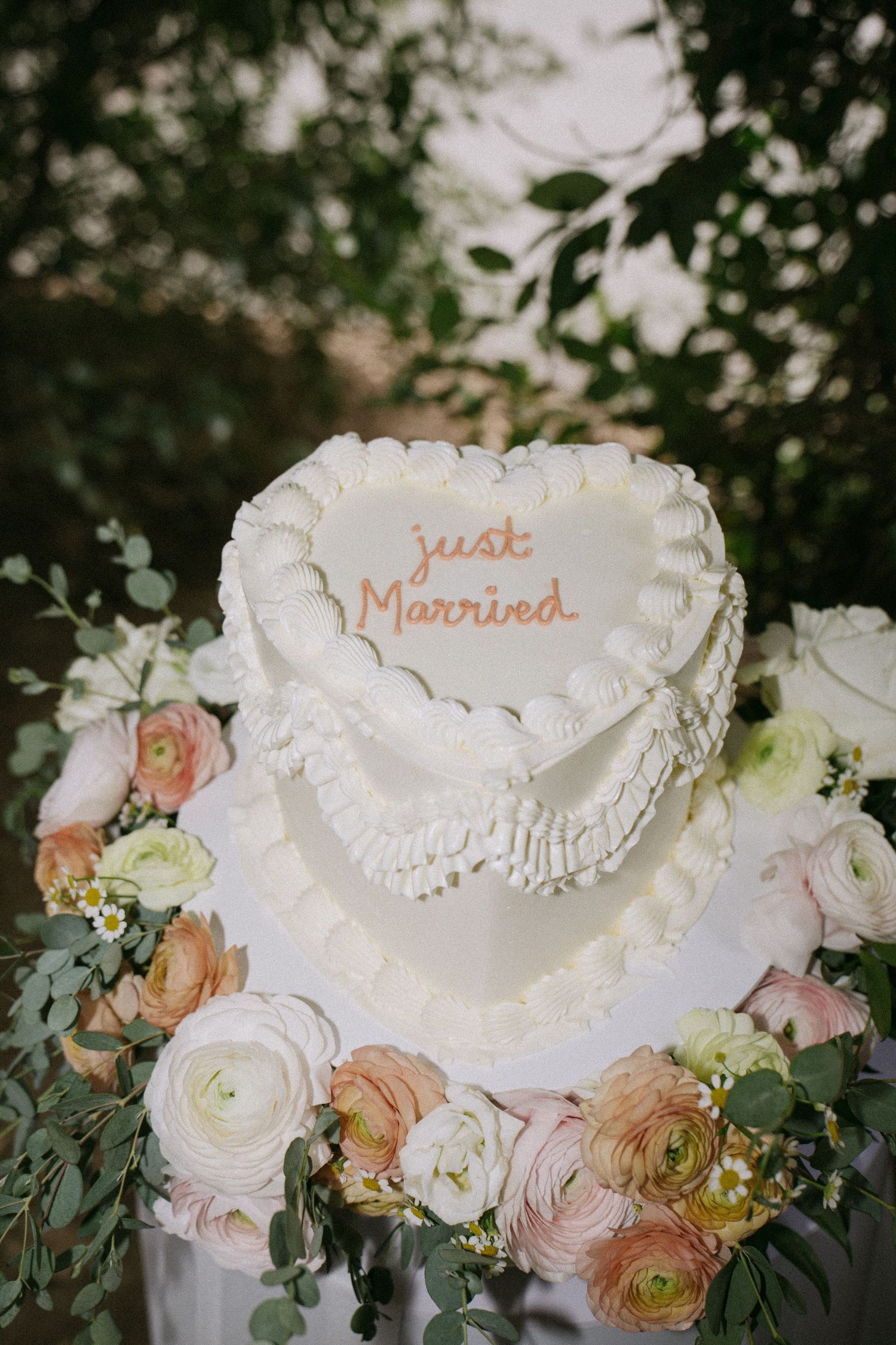 White heart-shaped two-tier wedding cake reading Just Married surrounded by pink ranunculus and eucalyptus flowers.