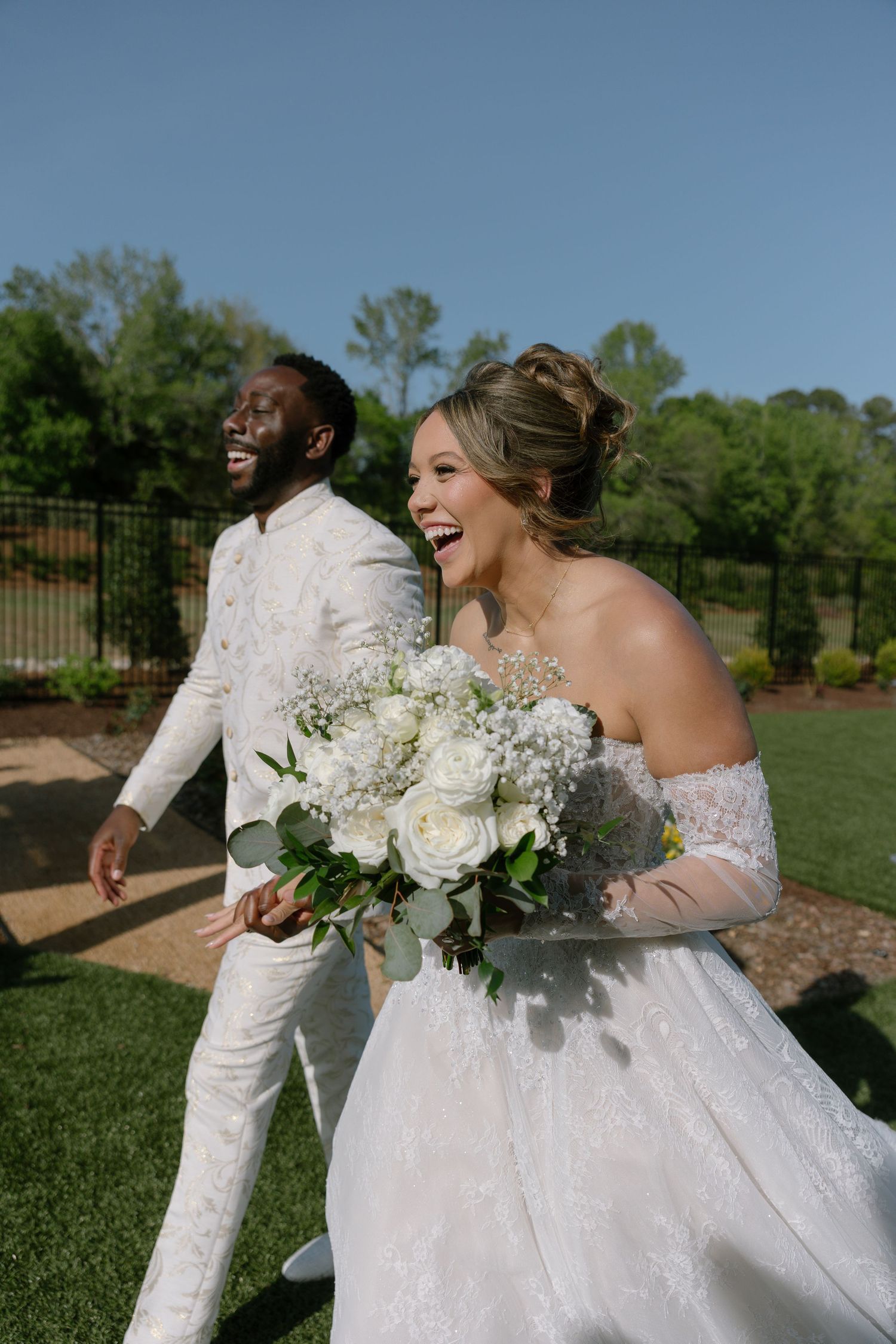 Joyful interracial couple in white wedding attire laughing outdoors, bride holding white rose bouquet on sunny day.