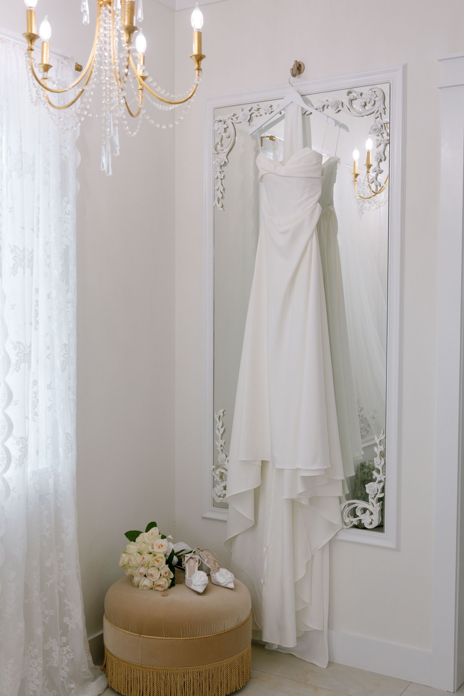 Elegant white wedding dress hanging on ornate mirror in bridal suite with chandelier and floral bouquet.