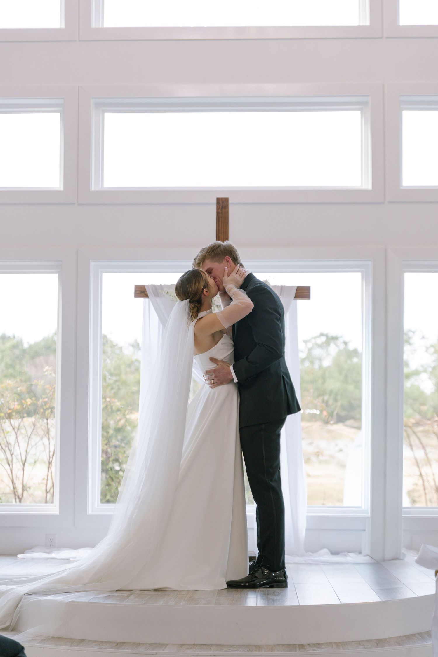Bride and groom share first kiss at altar before large windows with wooden cross in bright chapel.
