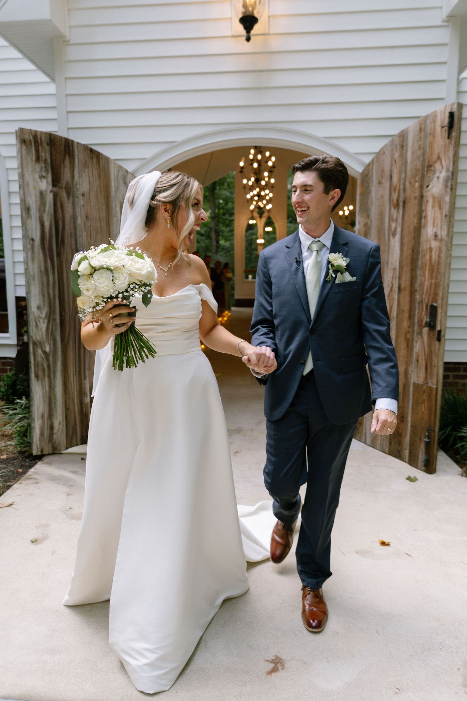 Bride and groom walk hand in hand from chapel doors, bride holding white bouquet in elegant off-shoulder gown.
